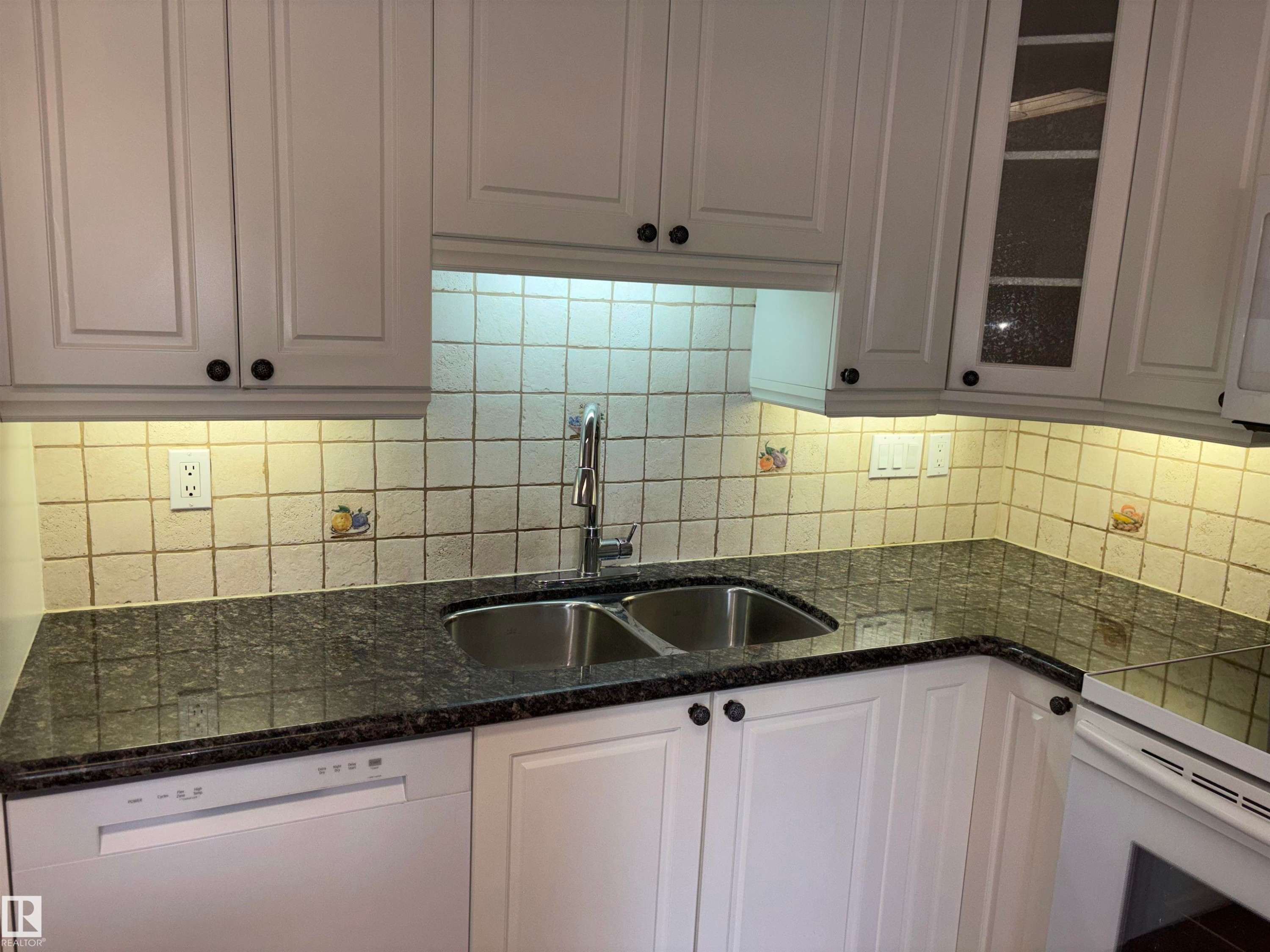 White cabinetry with black hardware, granite-style countertops, dual stainless steel sink, tiled backsplash, and under-cabinet lighting - 4027 74 Street Nw, Edmonton, AB