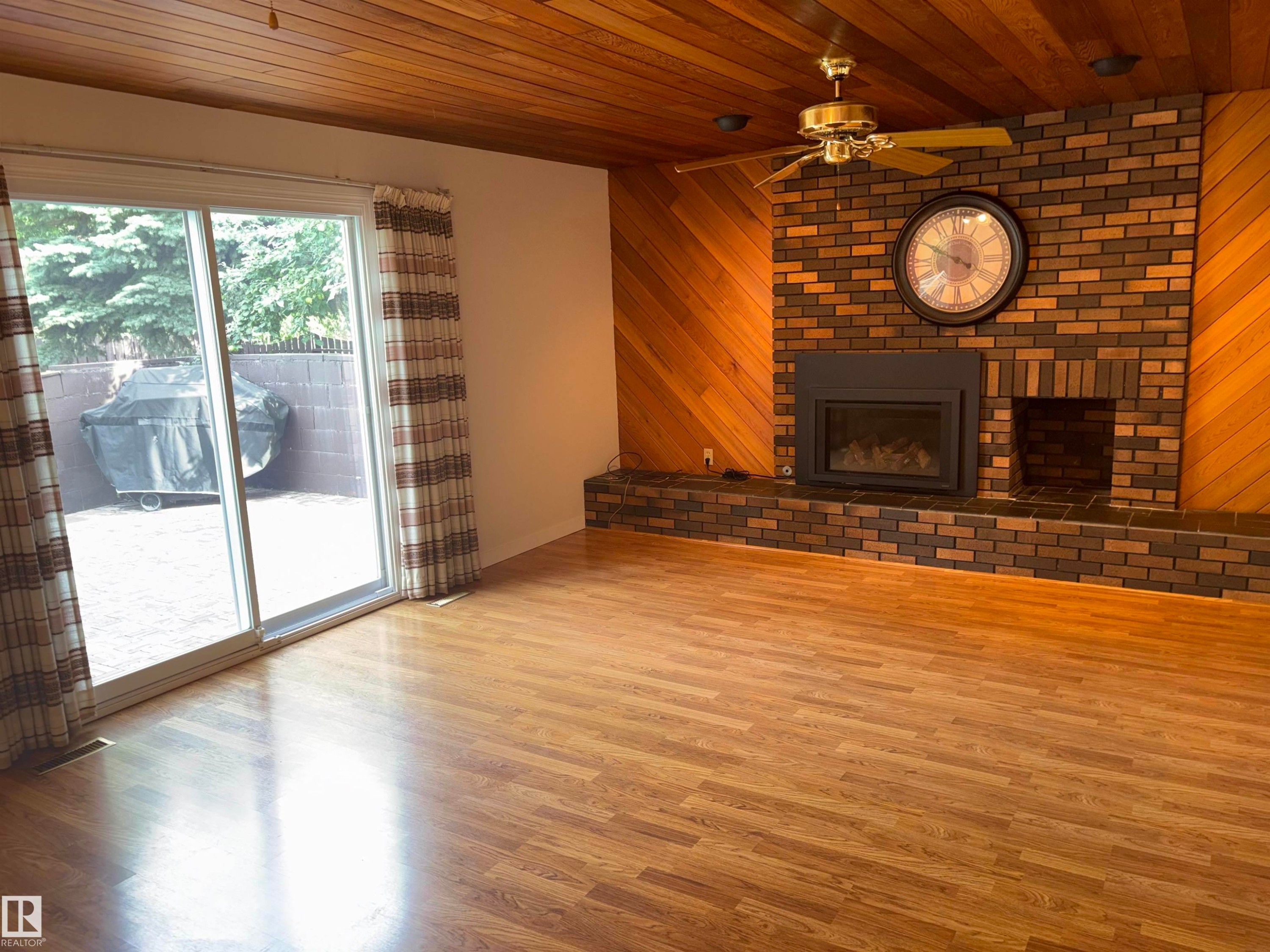 Wood-finish flooring, wood plank ceiling, brick fireplace with mantel, and sliding glass door to a paved patio - 4027 74 Street Nw, Edmonton, AB