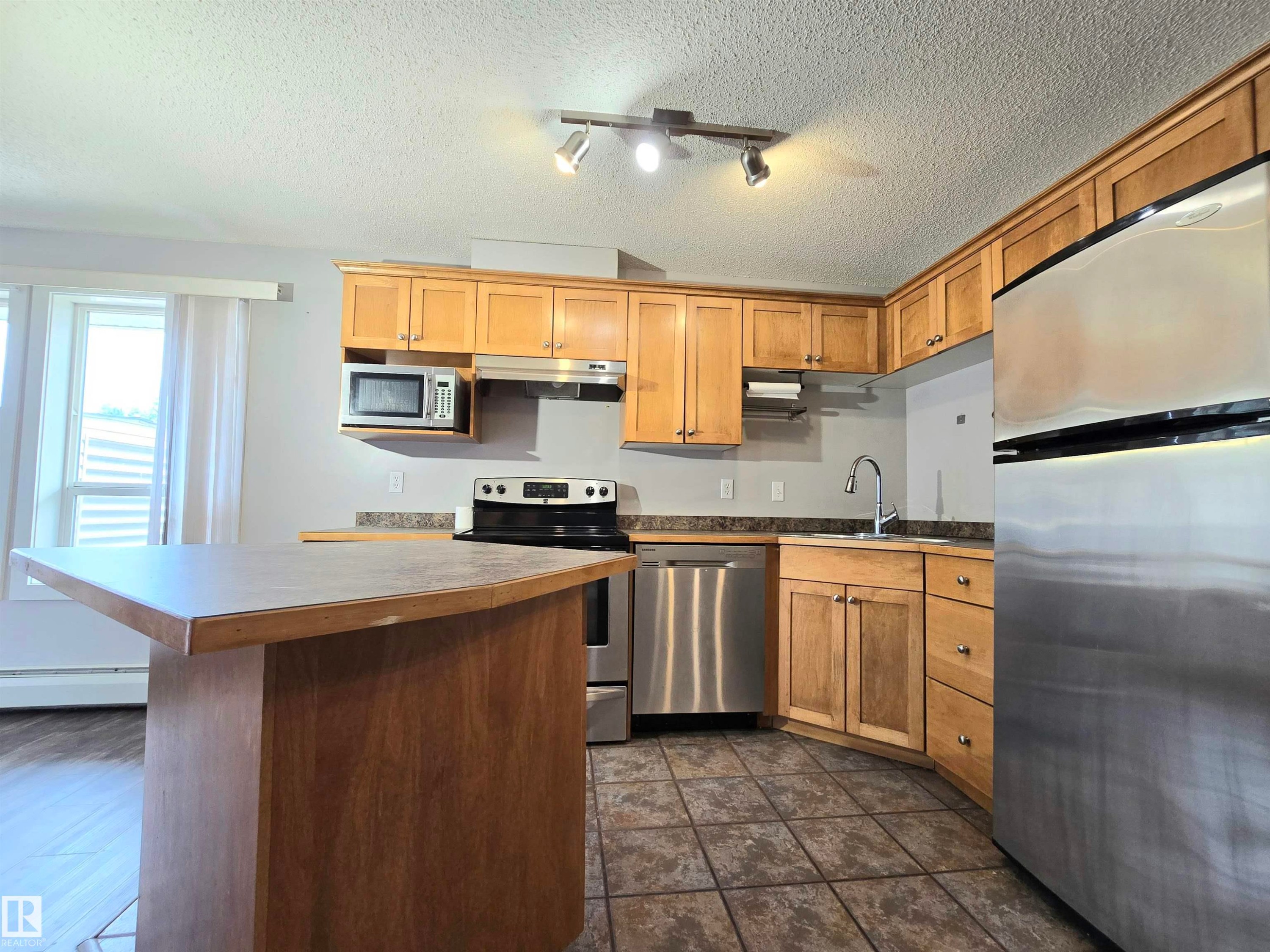 Kitchen featuring wood-finish cabinetry, stainless steel appliances, and a central island with an extended countertop - #105 155 Edwards Dr Sw, Edmonton, AB - Indoor Photo Showing Kitchen