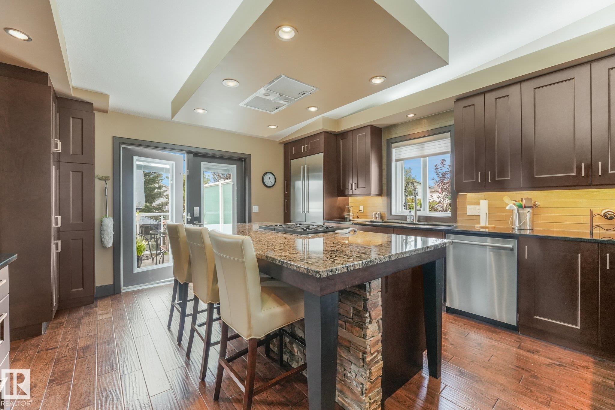 Kitchen featuring wood-finish flooring, dark wood cabinetry, a large central island with a stone slab countertop, a stack stone island base, and stainless steel appliances - 12827 150 Avenue Nw, Edmonton, AB
