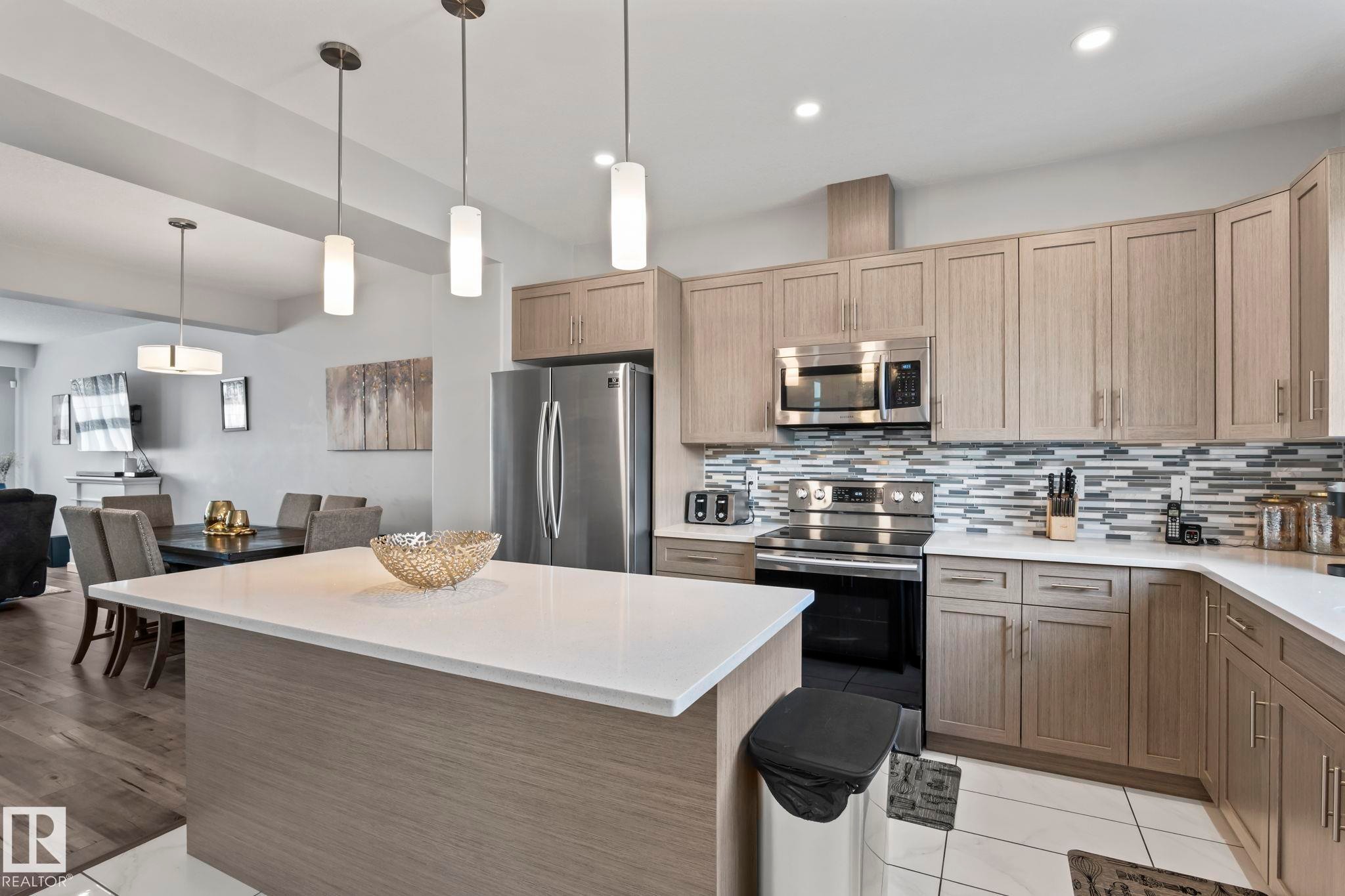 Open-concept kitchen with a central island, stainless steel appliances, light-toned cabinetry, and a patterned tile backsplash - 10338 162St Nw, Edmonton, AB