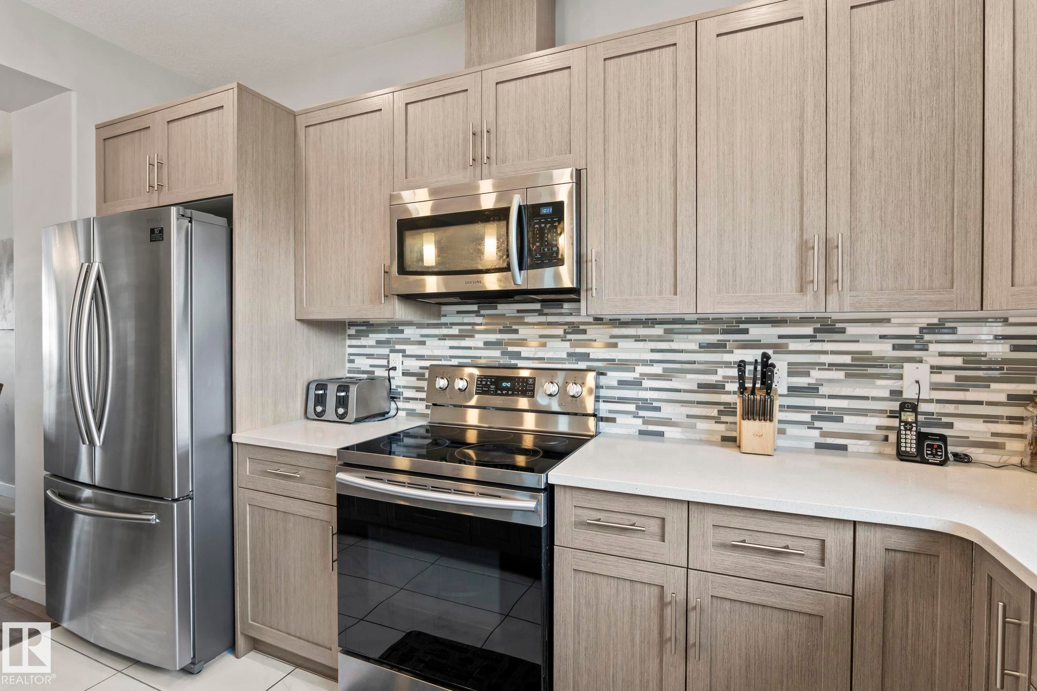 Kitchen featuring stainless steel appliances, light wood-finish cabinetry, a glass tile backsplash, and light-toned countertops - 10338 162St Nw, Edmonton, AB