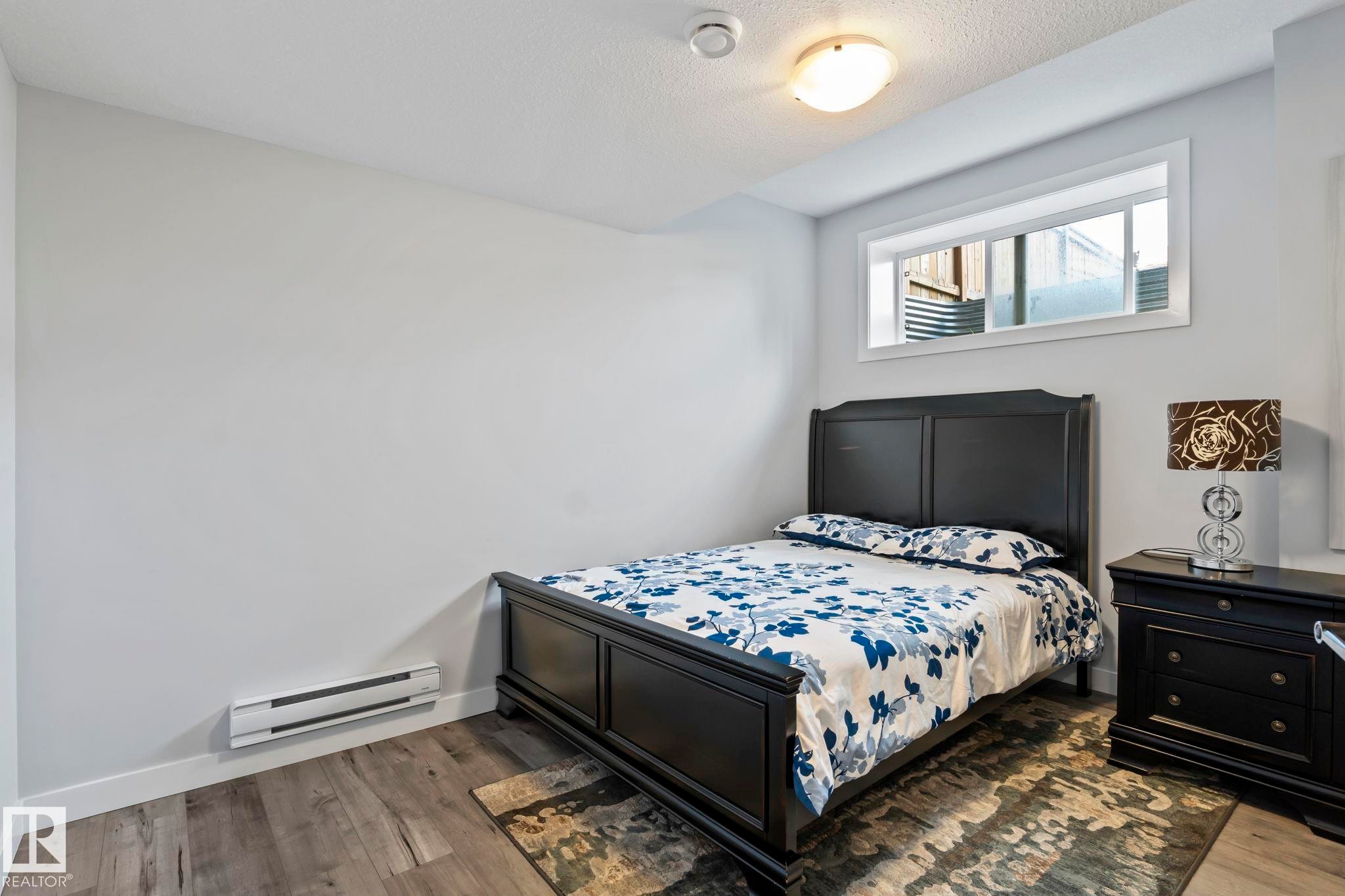 Window with white trim, wood-finish flooring, ceiling-mounted light fixture, and wall-mounted heating unit - 10338 162St Nw, Edmonton, AB