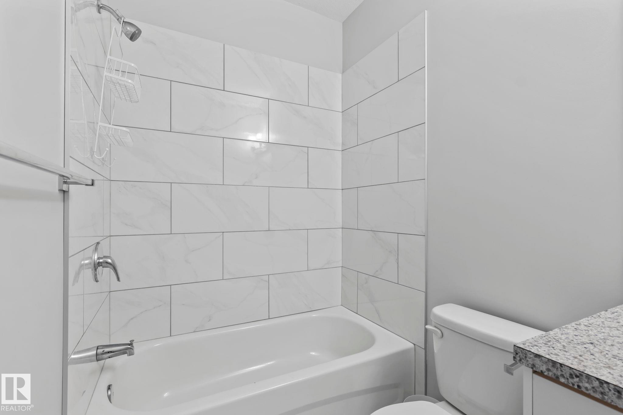 Bathtub with subway tile surround and chrome fixtures, adjacent to a toilet and vanity with a granite-style countertop - 10338 162St Nw, Edmonton, AB