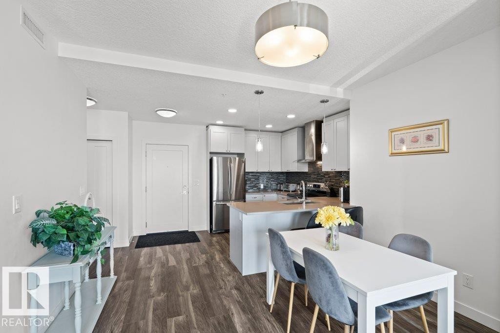 Open-concept kitchen and dining area featuring wood-finish flooring, white cabinetry, stainless steel appliances, and a dark stone-look backsplash - 1316 Windermere Way Sw, Edmonton, AB