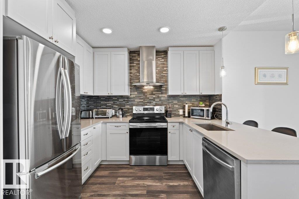 U-shaped kitchen featuring white cabinetry, stainless steel appliances, wood-finish flooring, a mosaic tile backsplash, and light-toned countertops - 1316 Windermere Way Sw, Edmonton, AB
