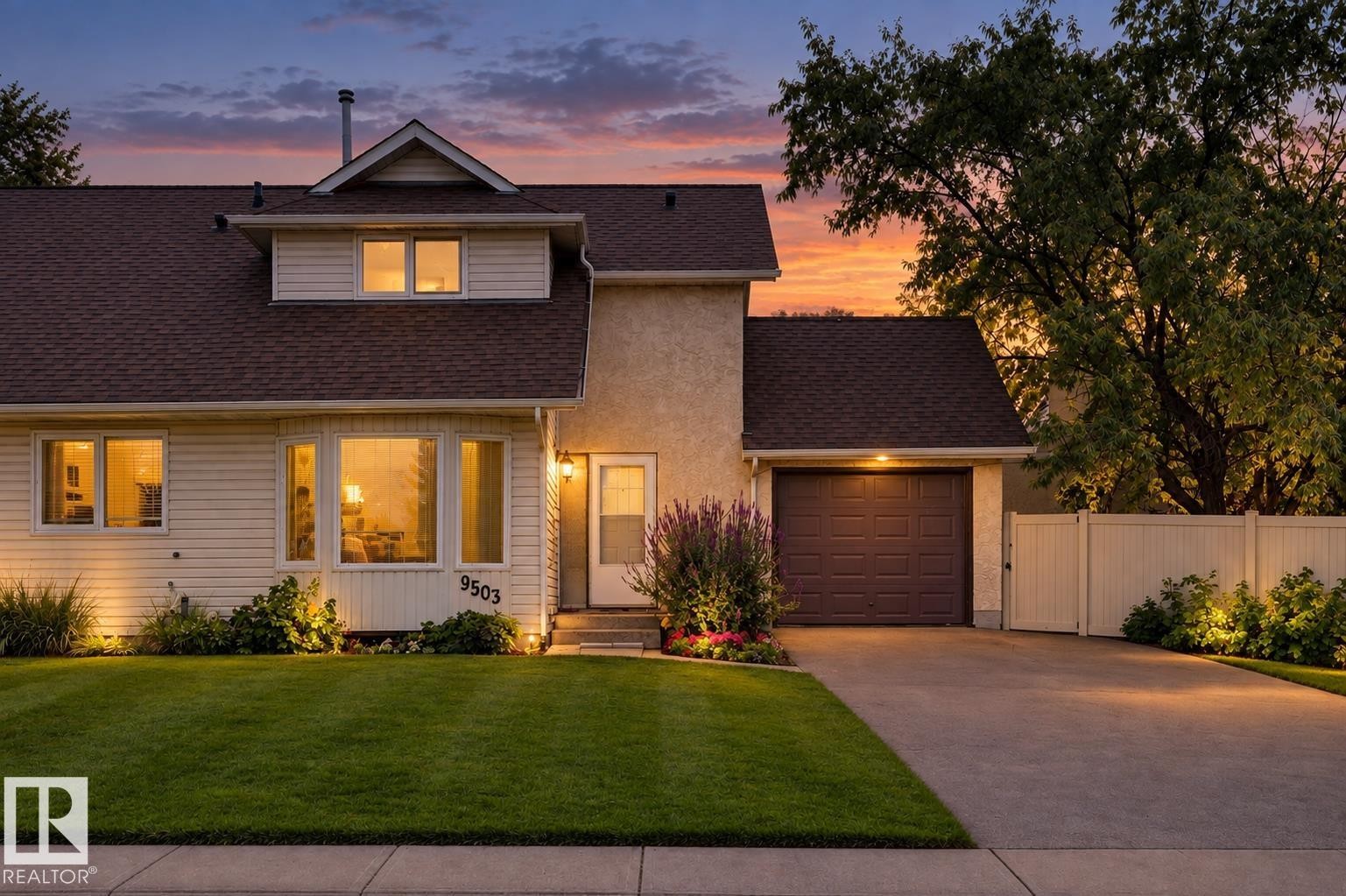 Exterior featuring light-colored siding and stucco, a two-car garage with a brown door, a front lawn, and a light-colored privacy fence - 9503 180A Street Nw, Edmonton, AB