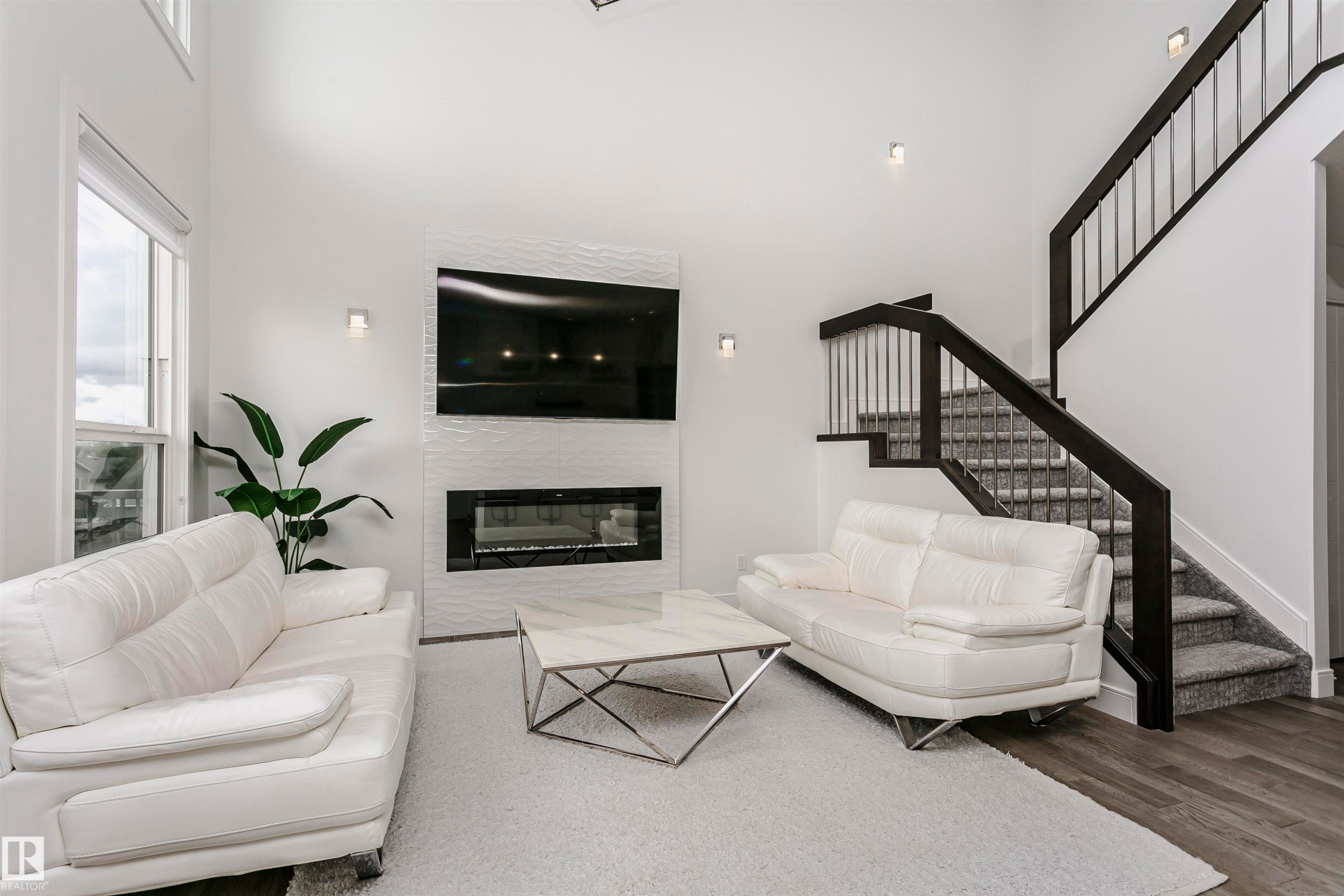 Two-story living area featuring a modern fireplace with textured tile surround, wood-finish flooring, and a contemporary staircase with dark wood railings and metal spindles - 9107 181 Avenue Nw, Edmonton, AB