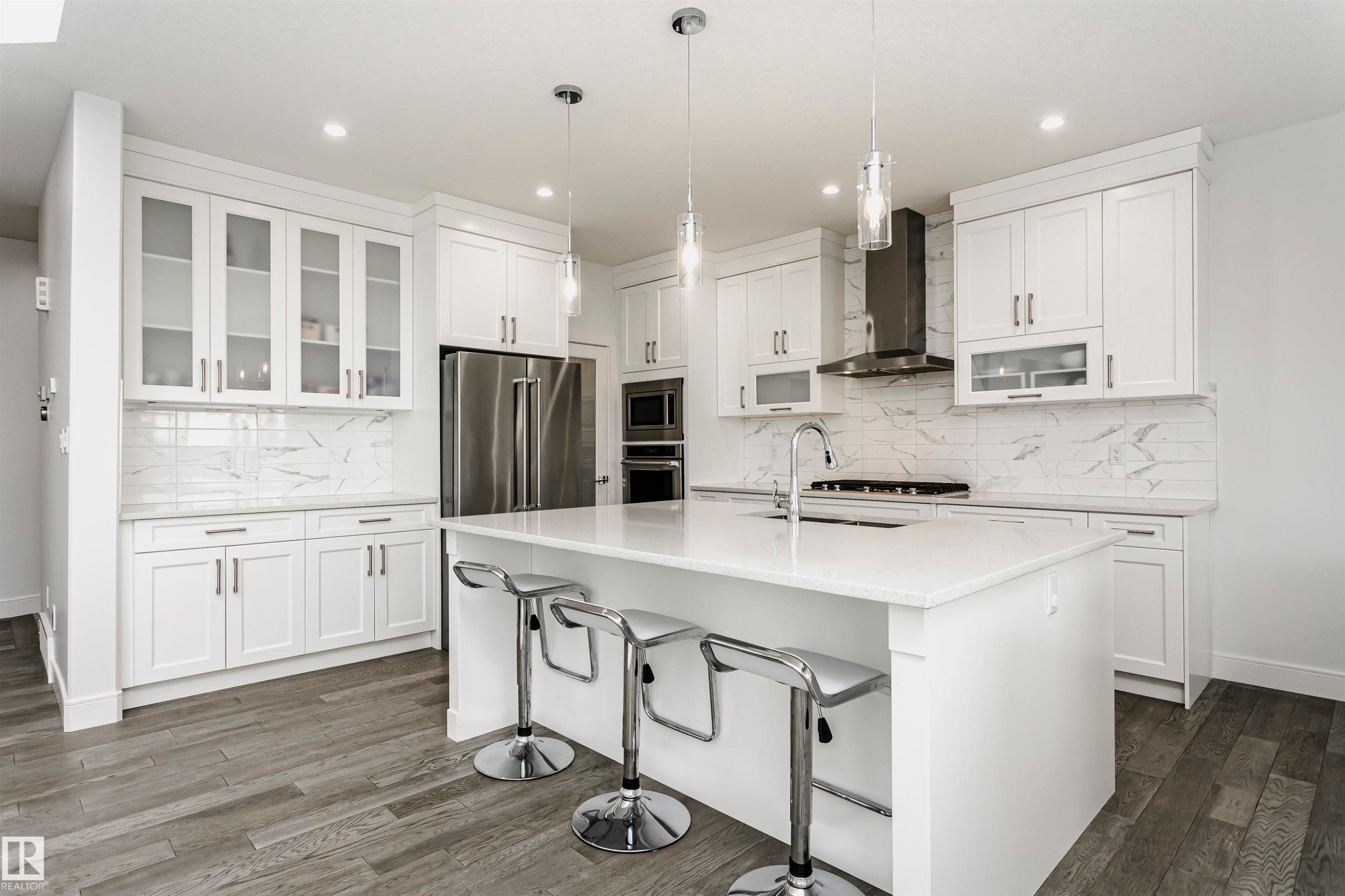 Contemporary white cabinetry with glass-front accents, white countertop island, stainless steel appliances, and wood-finish flooring - 9107 181 Avenue Nw, Edmonton, AB