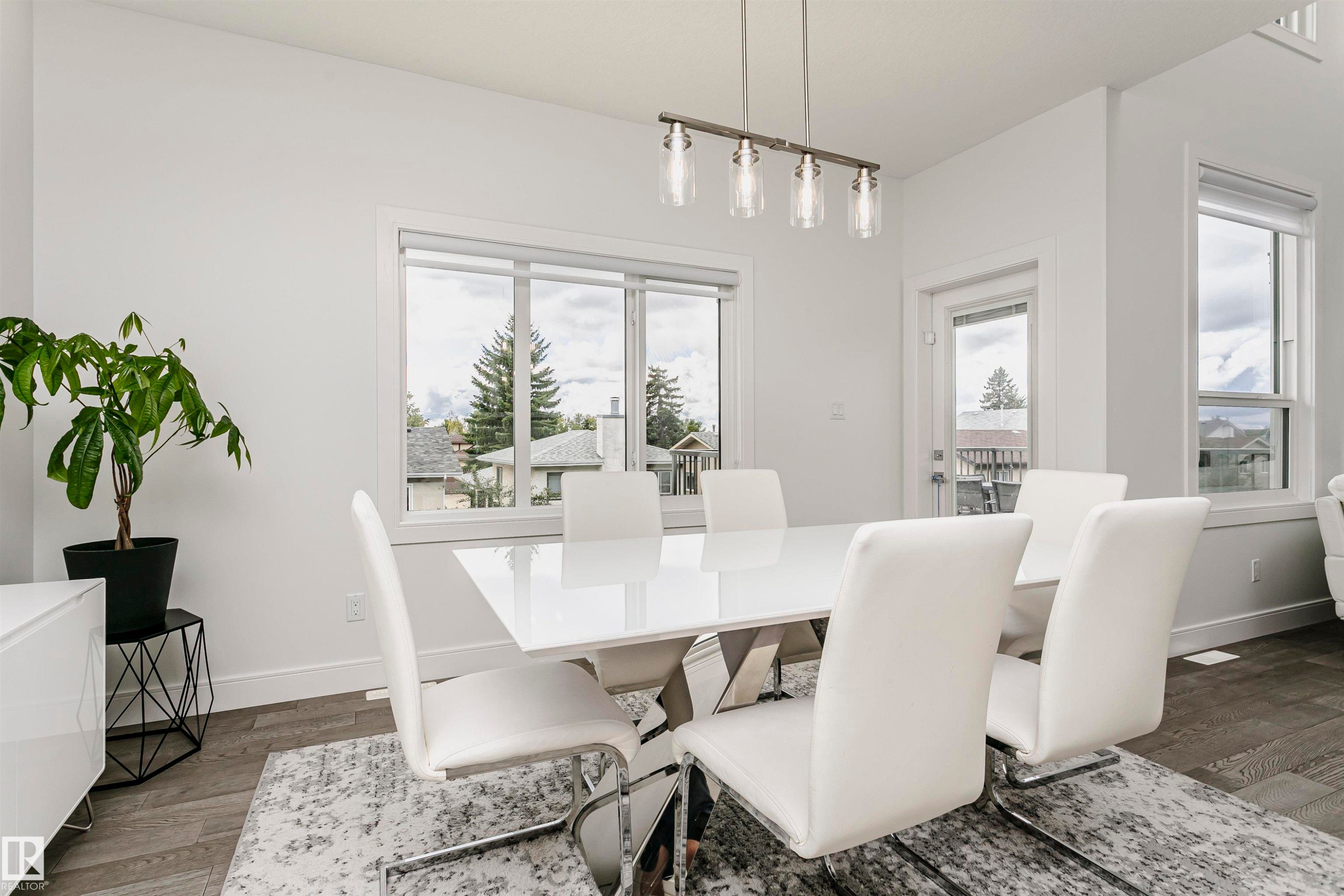 Open-concept dining area featuring wood-finish flooring, a modern chandelier with clear glass shades, large windows with white trim, and a glass-paneled exterior door - 9107 181 Avenue Nw, Edmonton, AB