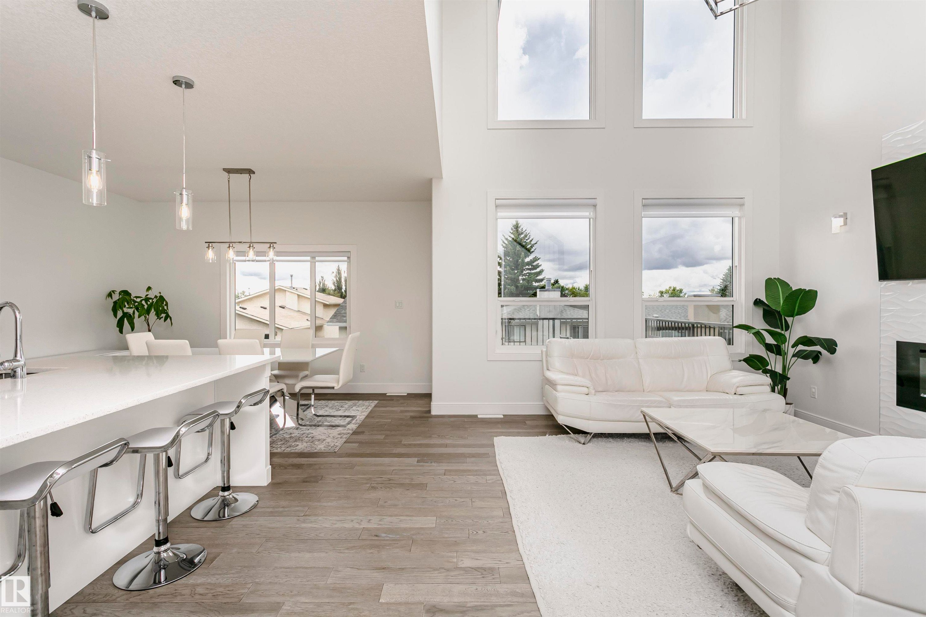 Open-concept living area with soaring ceilings, wood-finish flooring, a contemporary fireplace, and an expansive kitchen island with a white countertop - 9107 181 Avenue Nw, Edmonton, AB