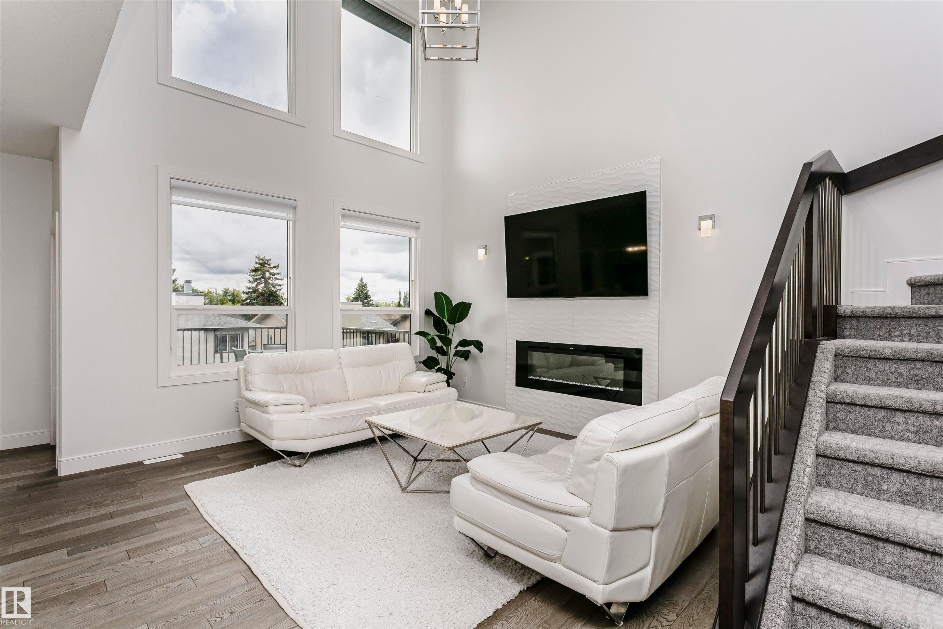 Living area featuring a modern linear fireplace with a textured tile surround, high ceilings, large windows, wood-finish flooring, and a dark wood staircase with carpeted treads - 9107 181 Avenue Nw, Edmonton, AB