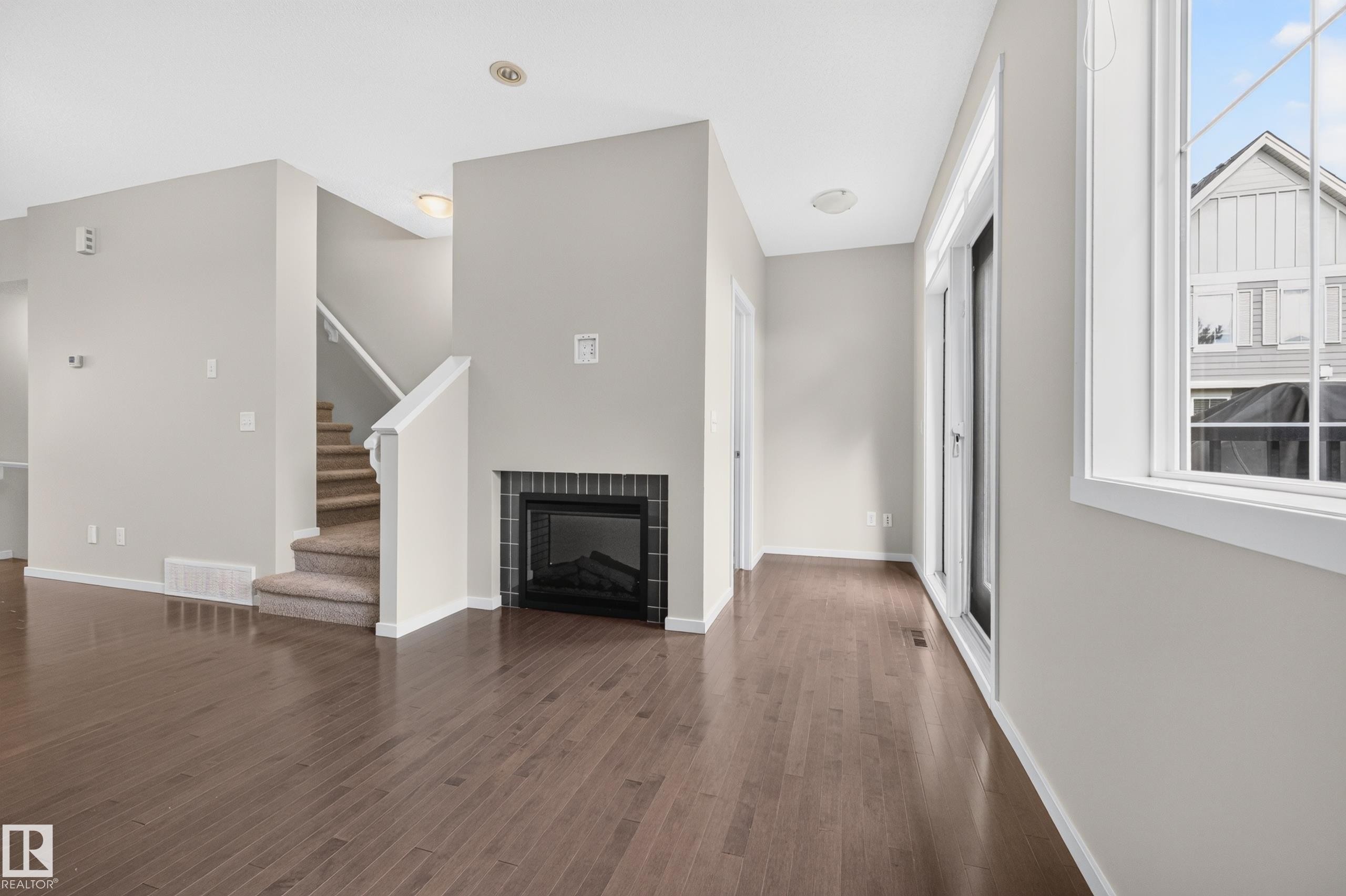 Wood-finish flooring throughout, a modern fireplace with a dark tile surround, a staircase with white balusters and carpeted treads, and abundant natural light from multiple windows - #72 603 Watt Bv Sw Sw, Edmonton, AB