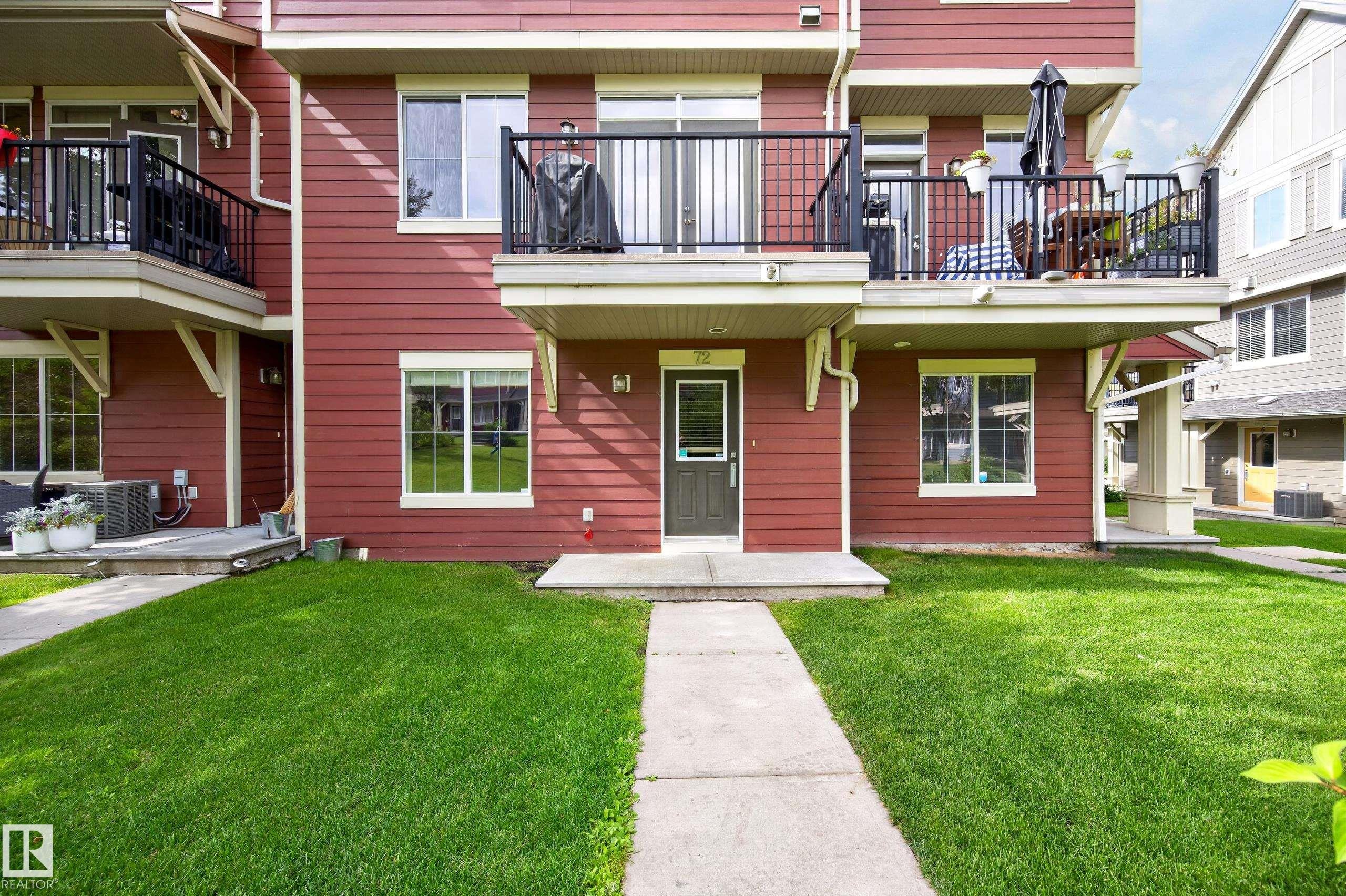 Red horizontal siding exterior with a dark gray entry door, concrete walkway, ground-level windows, and an upper-level balcony with black railings - #72 603 Watt Bv Sw Sw, Edmonton, AB