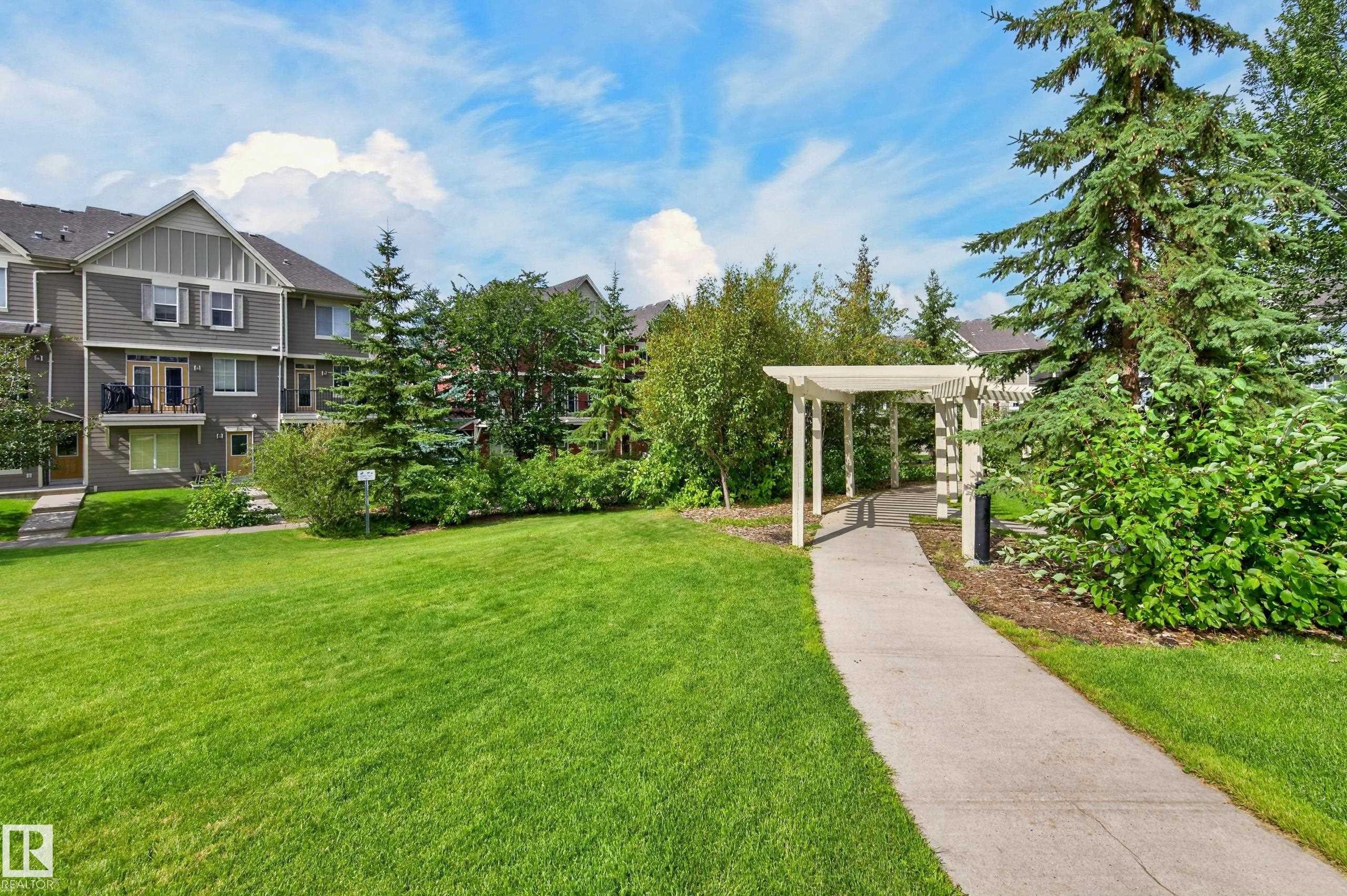 Expansive green space featuring a concrete pathway, white pergola, and mature trees - #72 603 Watt Bv Sw Sw, Edmonton, AB