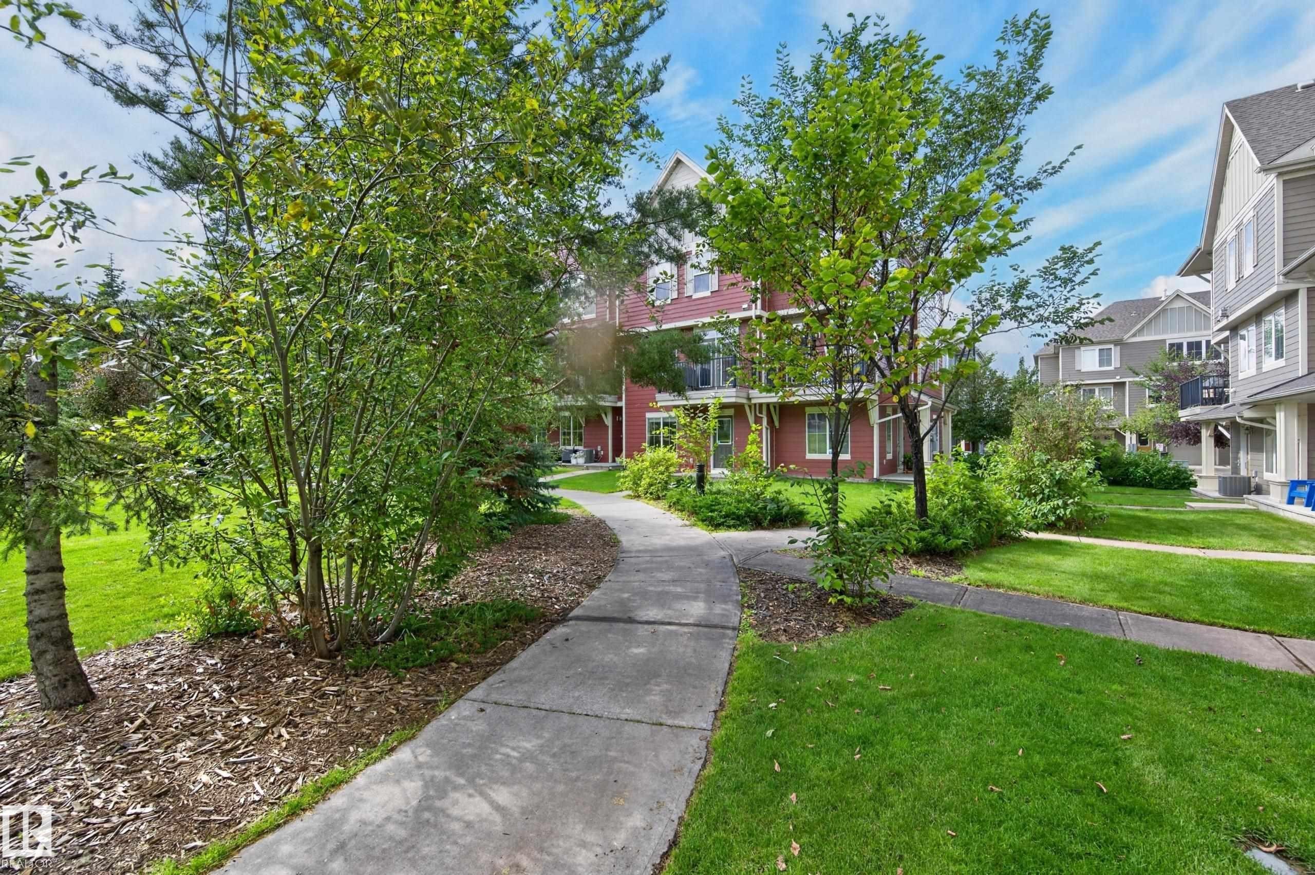 Landscaped grounds featuring a winding concrete pathway, mature trees, and shrubs - #72 603 Watt Bv Sw Sw, Edmonton, AB