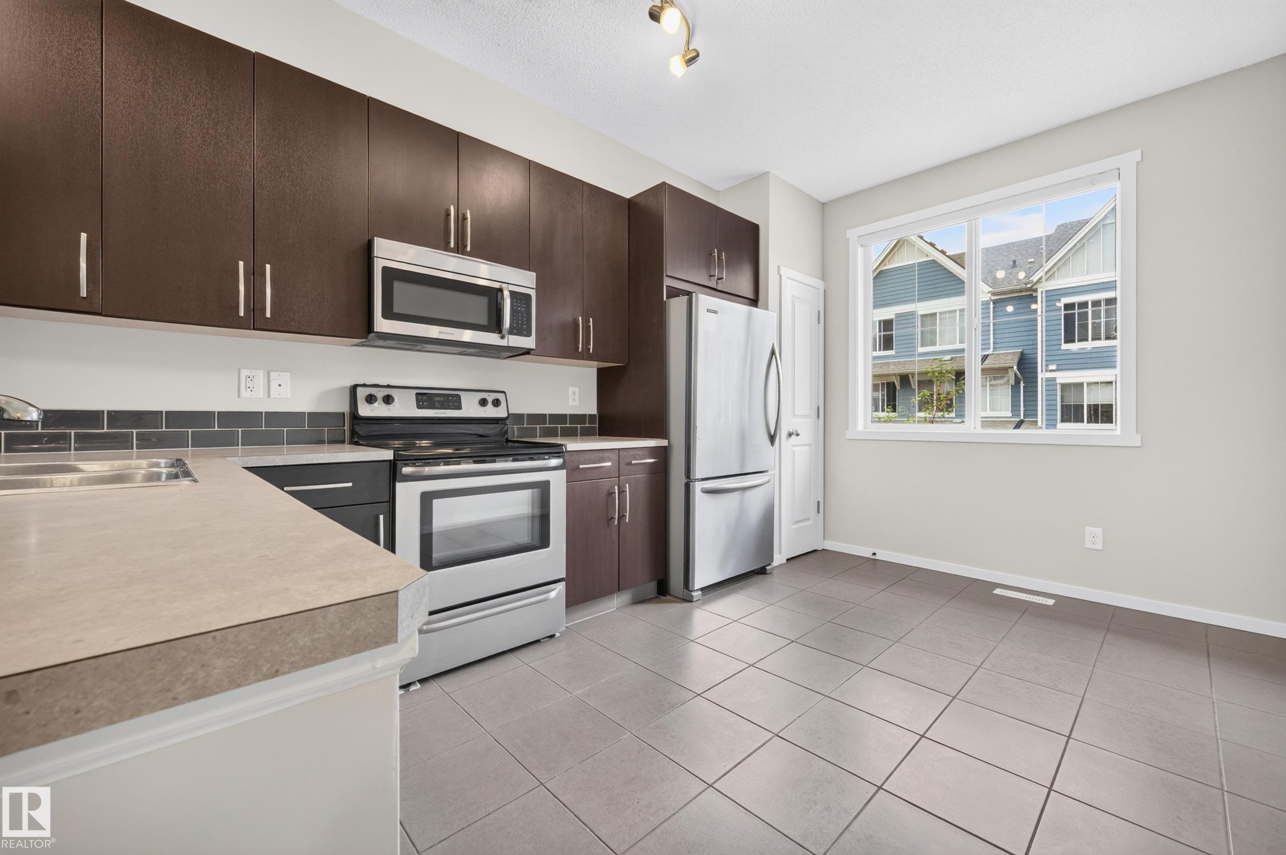 Kitchen featuring dark wood-finish cabinetry, stainless steel appliances, light-toned countertops, a subway tile backsplash, and ceramic tile flooring - #72 603 Watt Bv Sw Sw, Edmonton, AB