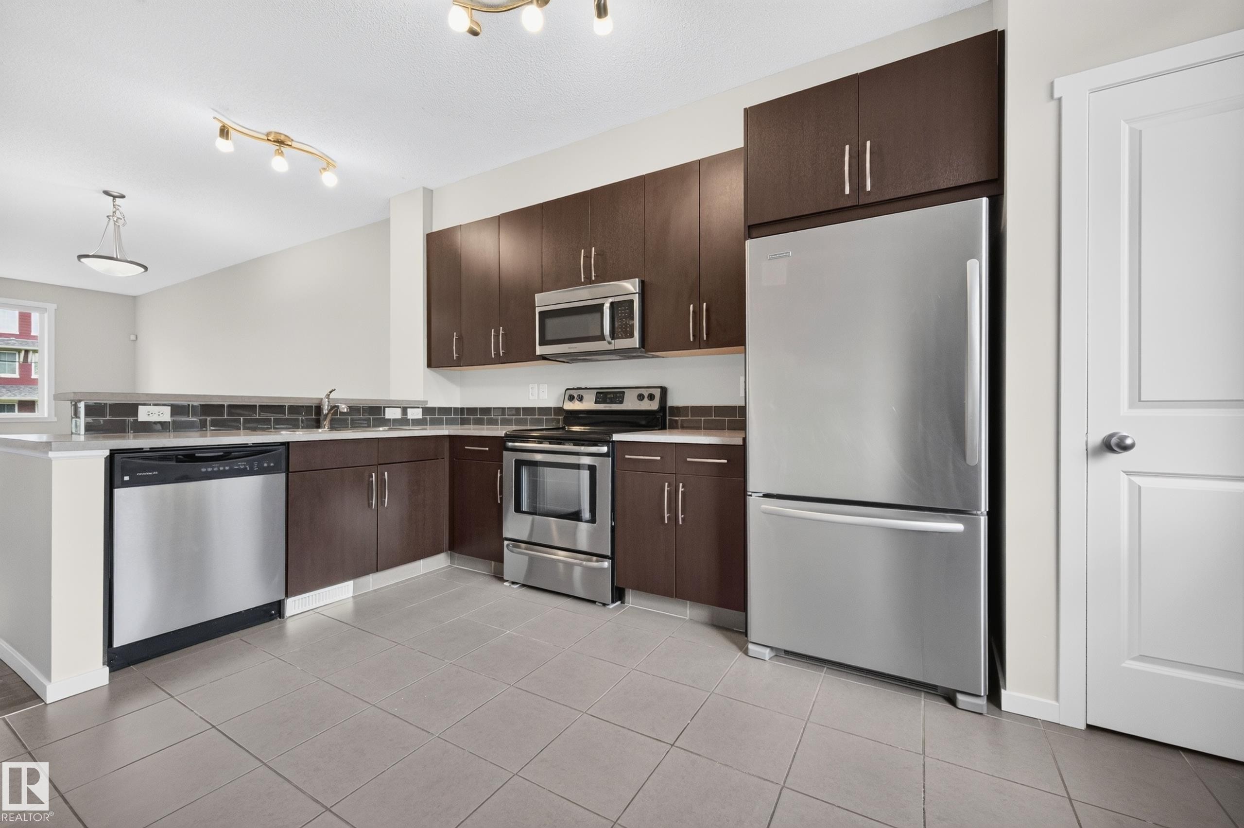 Kitchen featuring wood-finish cabinetry, stainless steel appliances, a subway tile backsplash, and light gray floor tiling - #72 603 Watt Bv Sw Sw, Edmonton, AB