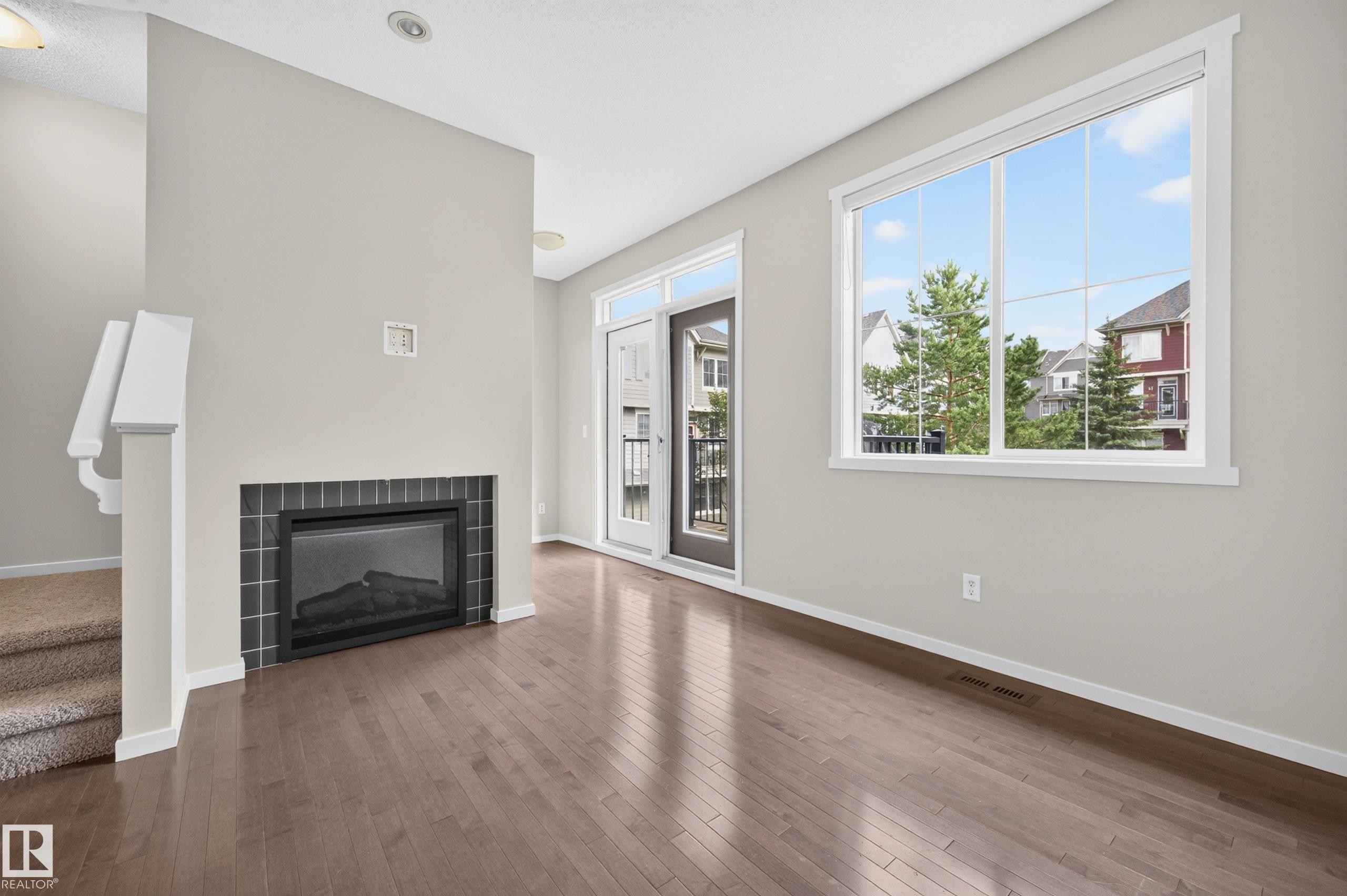 Wood-finish flooring, built-in fireplace with dark tile surround, white trim, sliding glass door with balcony, and large window - #72 603 Watt Bv Sw Sw, Edmonton, AB