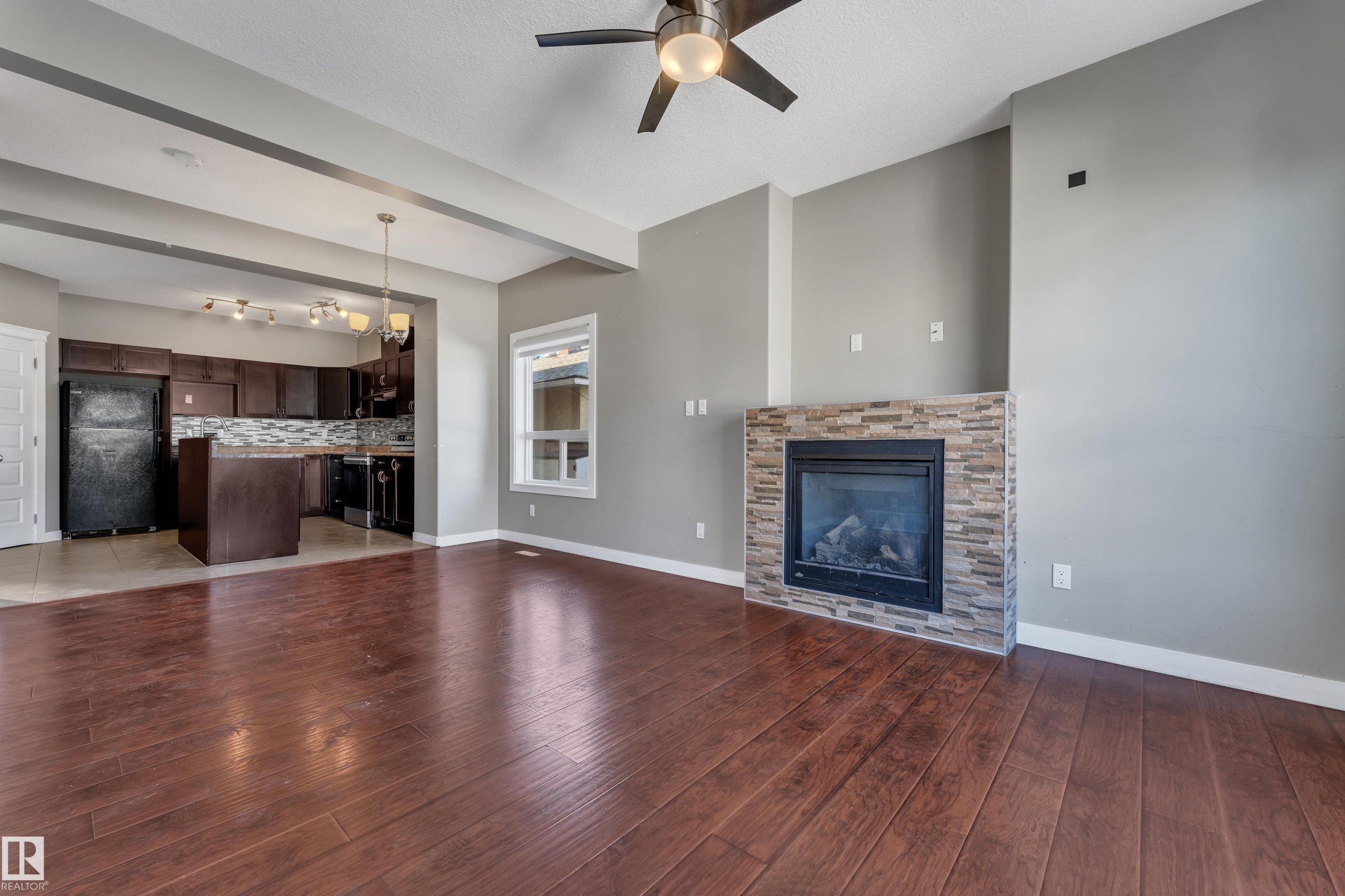Stone-clad fireplace feature wall, wood-finish flooring, kitchen with dark wood cabinetry, and a ceiling fan with integrated lighting - 11421 82 Street Nw, Edmonton, AB