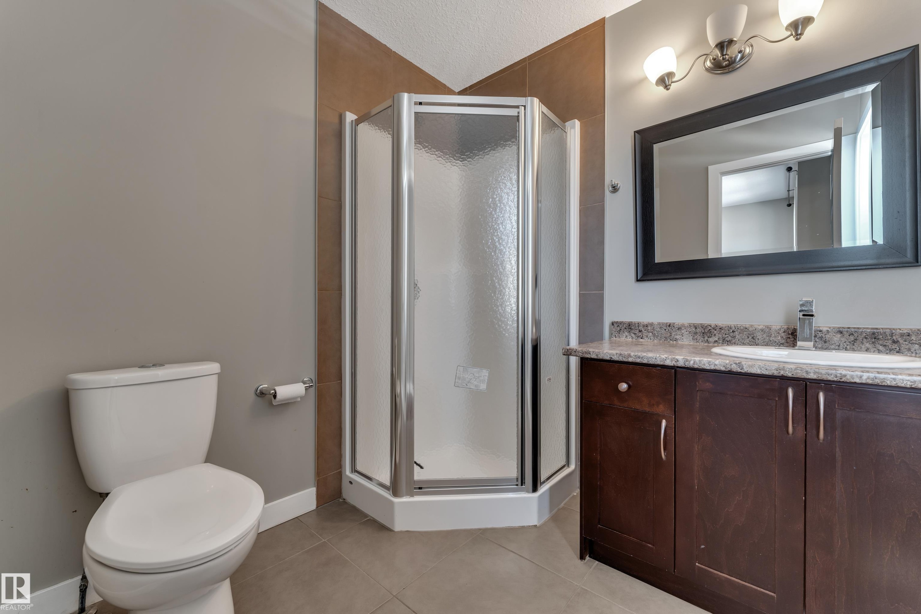 Corner shower with a glass enclosure, single vanity with a stone countertop, dark wood-finish cabinetry, and a framed mirror - 11421 82 Street Nw, Edmonton, AB