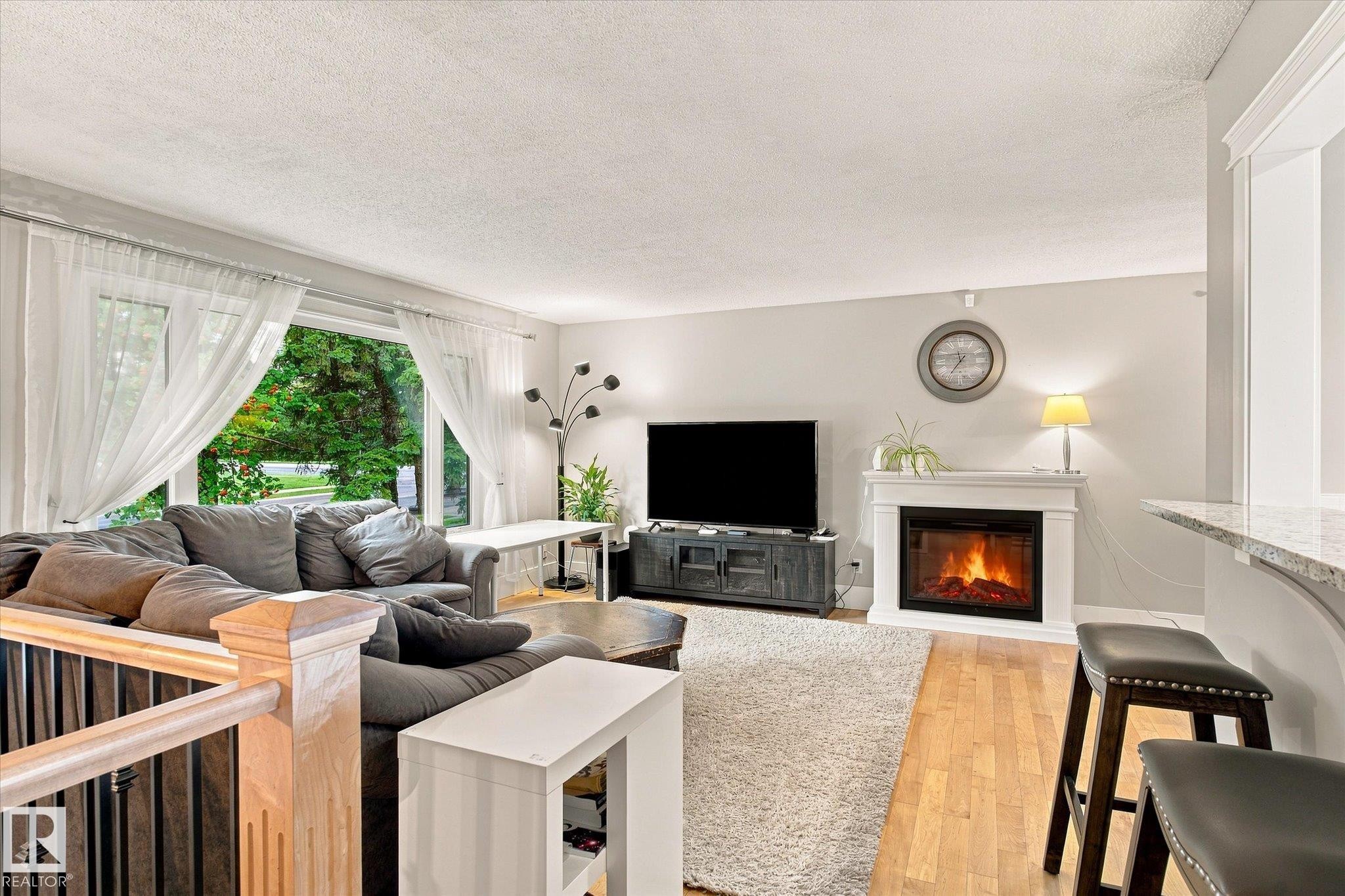 Hardwood-style flooring throughout, featuring a white mantle fireplace with hearth, an integrated breakfast bar with a stone-finish countertop, a large picture window, and a wood-finish staircase with black spindles - 3 Akins Drive, St. Albert, AB