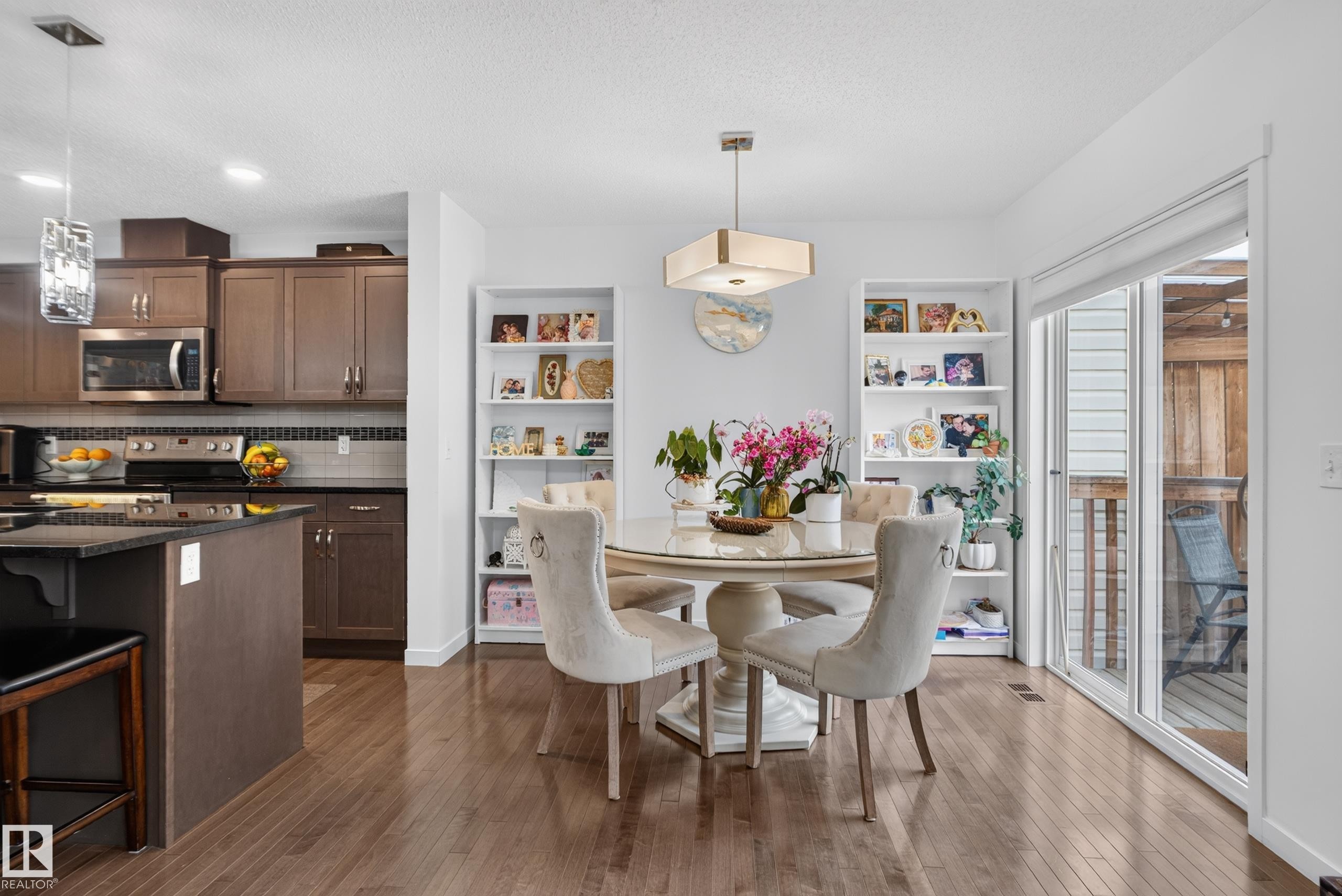 Wood-finish flooring throughout, kitchen with dark cabinetry and stainless steel appliances, built-in shelving, dining area with a modern chandelier, and a sliding glass door - 17363 120 Street Nw, Edmonton, AB