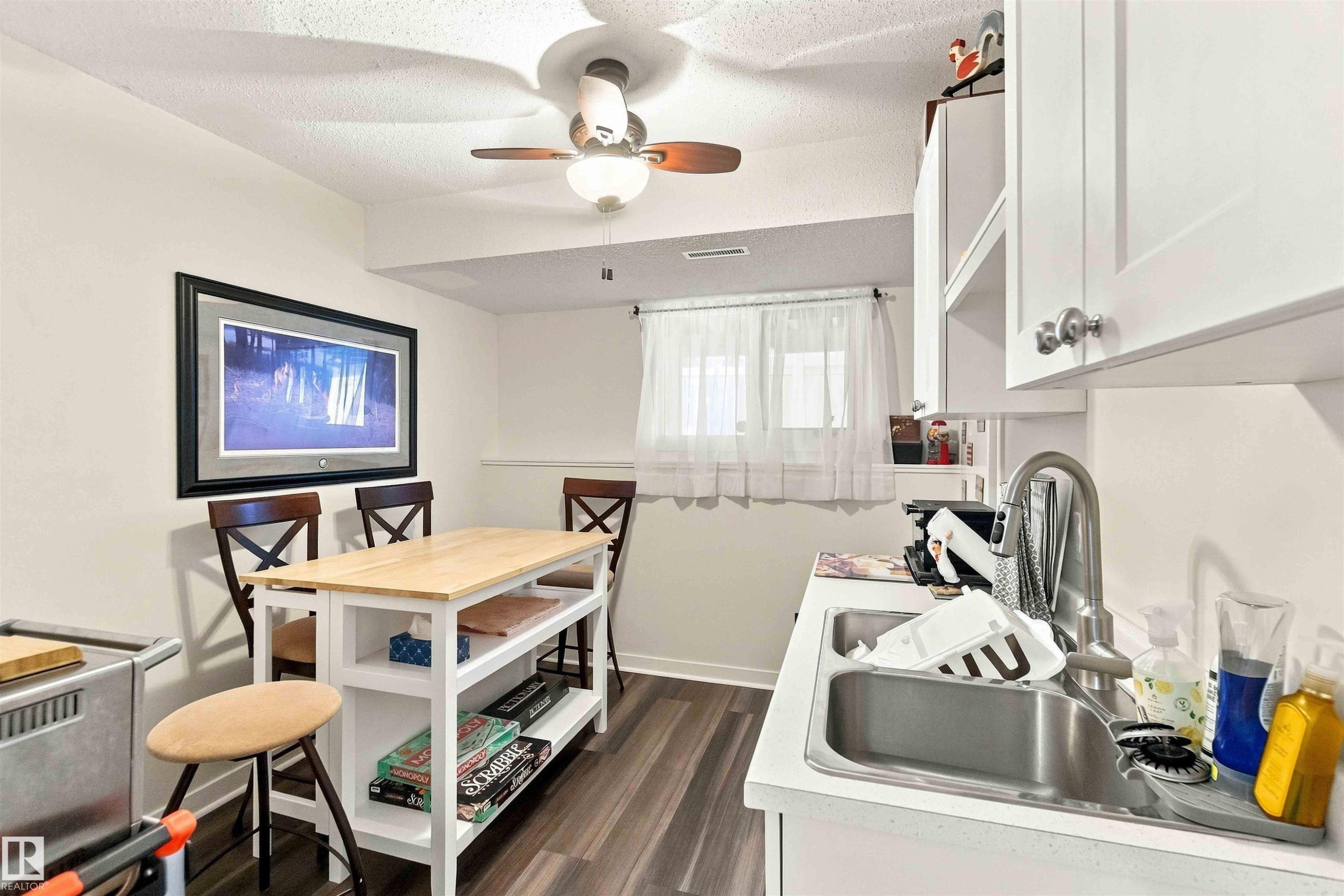 Efficient kitchen layout featuring stainless steel sink with faucet, white cabinetry with upper storage, wood-finish flooring, and adjacent dining area with seating for four - #6 15 Poirier Av, St. Albert, AB