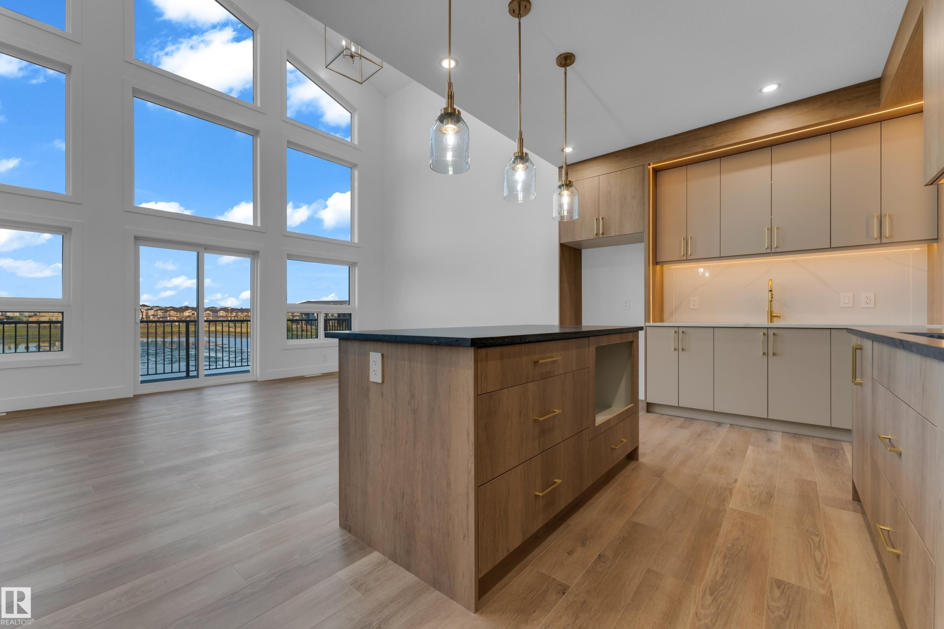 Soaring vaulted ceilings with expansive windows, contemporary kitchen island with dark countertop, elegant gold-tone hardware, wood-finish flooring, and modern pendant lighting - 236 Linden Loop, Leduc, AB