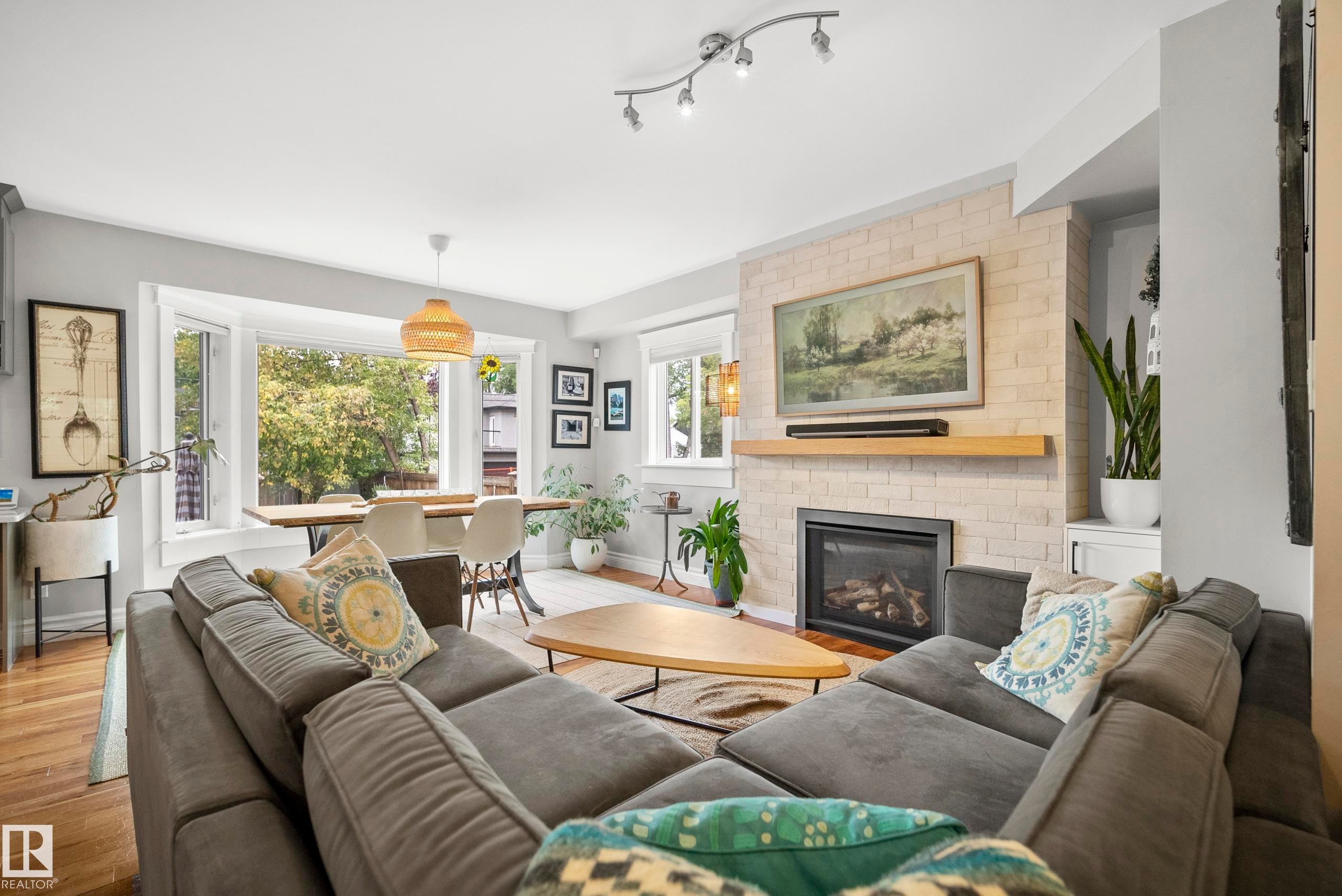Living area with wood-finish flooring, a contemporary fireplace featuring a light brick surround and wood mantle, and track lighting - 11006 64 Avenue Nw, Edmonton, AB