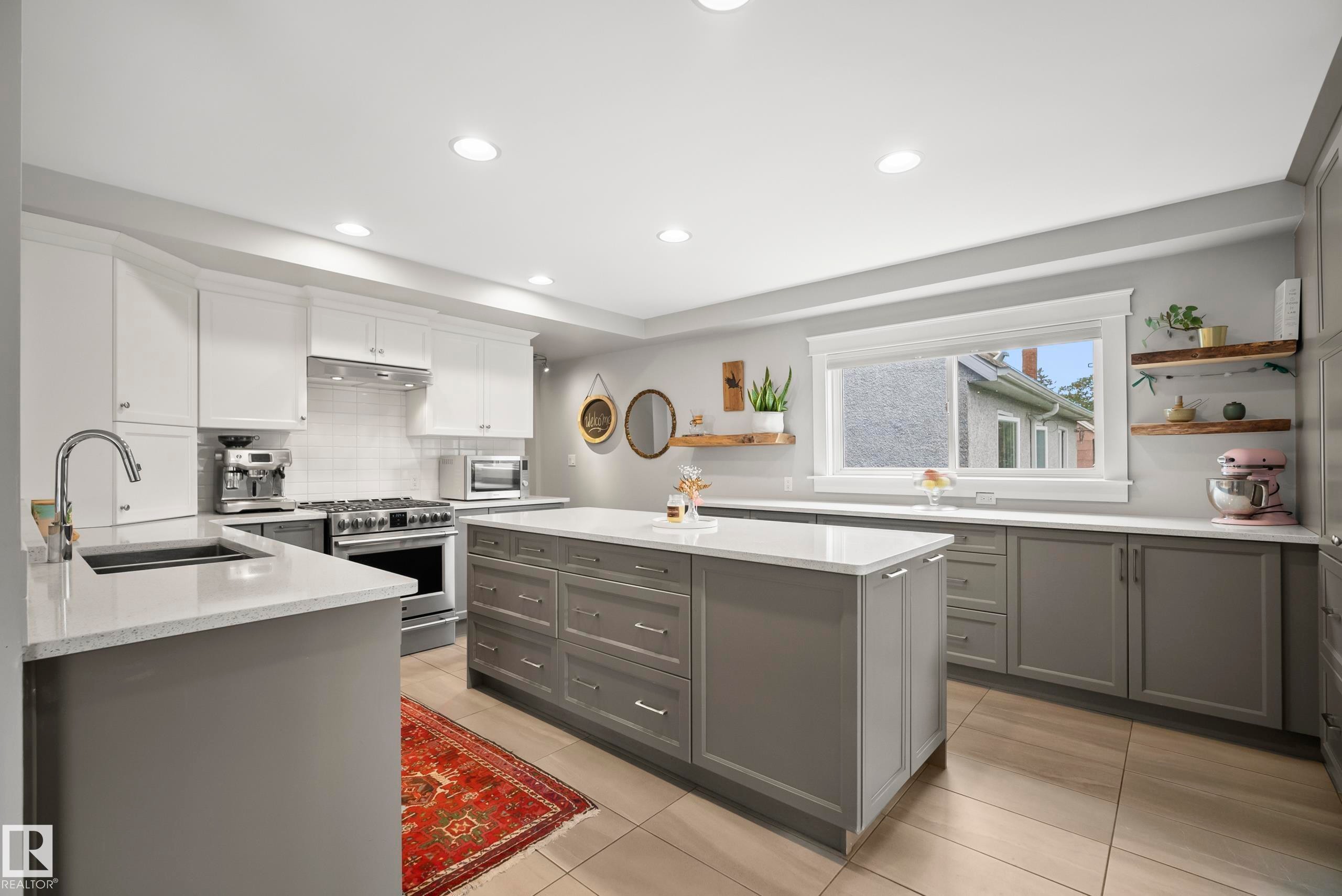 Modern kitchen featuring a central island with integrated storage, light-toned countertops, contrasting grey and white cabinetry, recessed lighting, and a stainless steel range with matching hood - 11006 64 Avenue Nw, Edmonton, AB