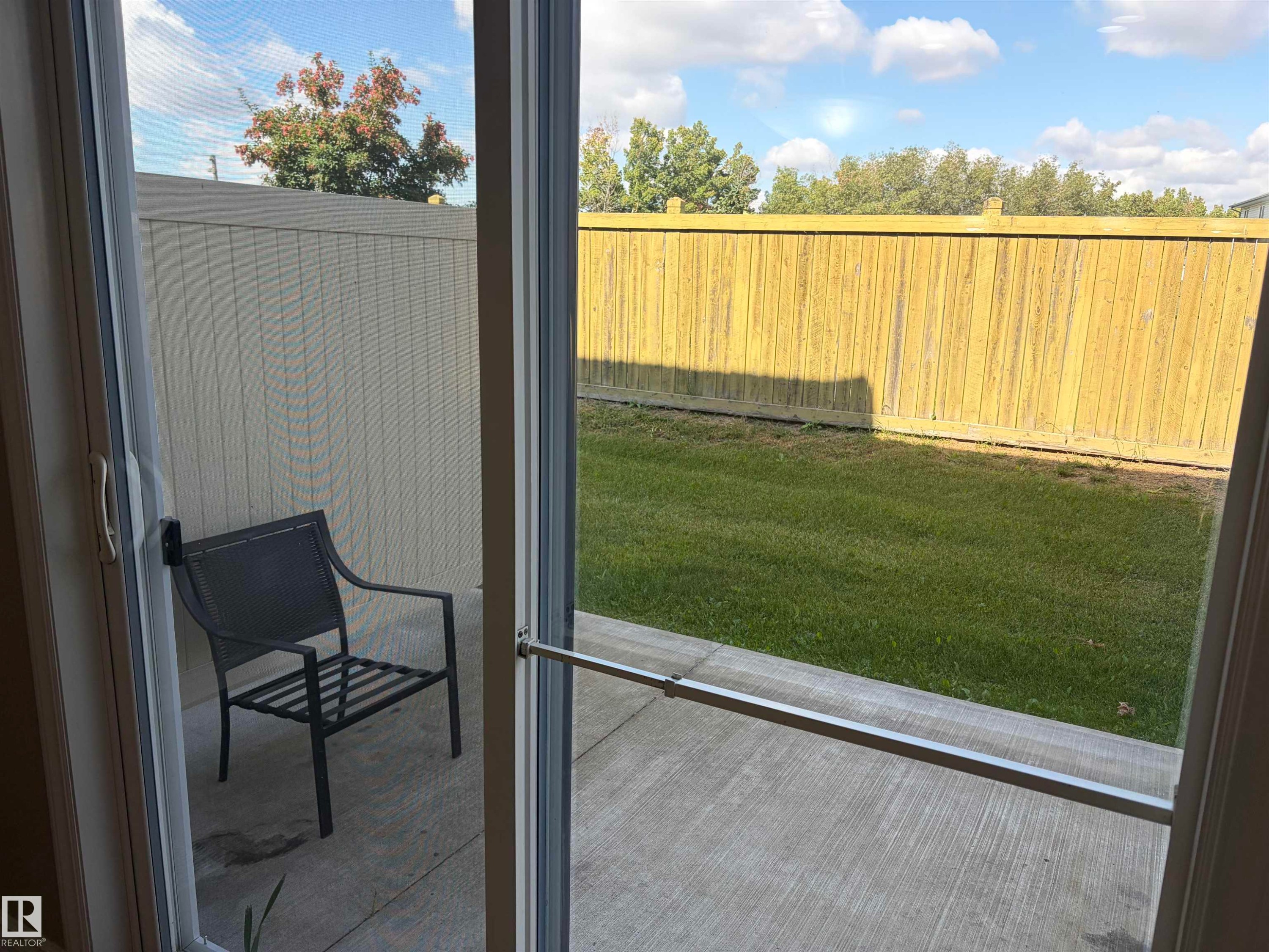 Sliding glass patio door with a screen, opening to a concrete patio and a fenced backyard with a manicured lawn - #100 13825 155 Av Nw, Edmonton, AB