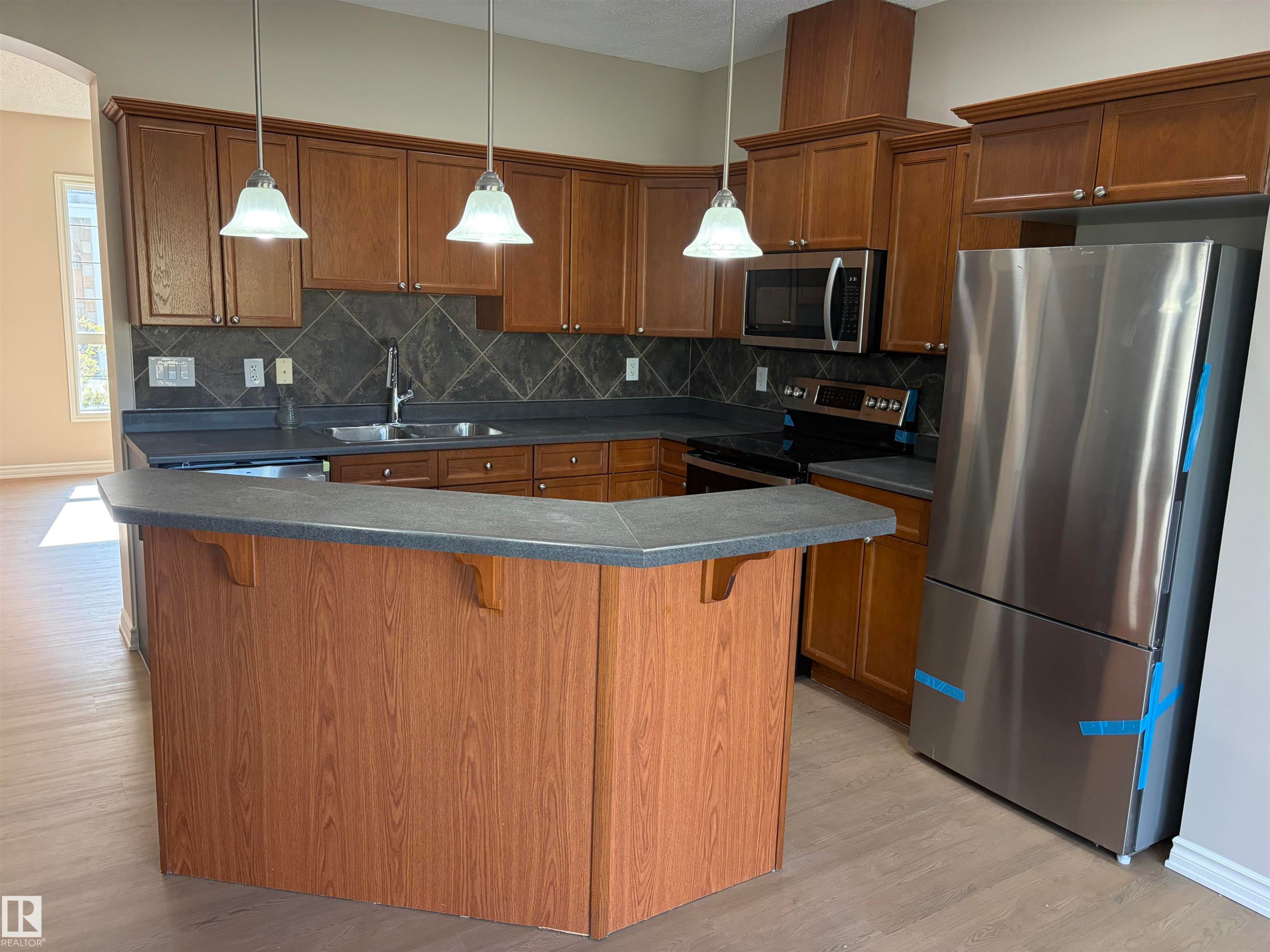 Kitchen featuring wood-finish cabinetry, dark countertops, and stainless steel appliances - #100 13825 155 Av Nw, Edmonton, AB