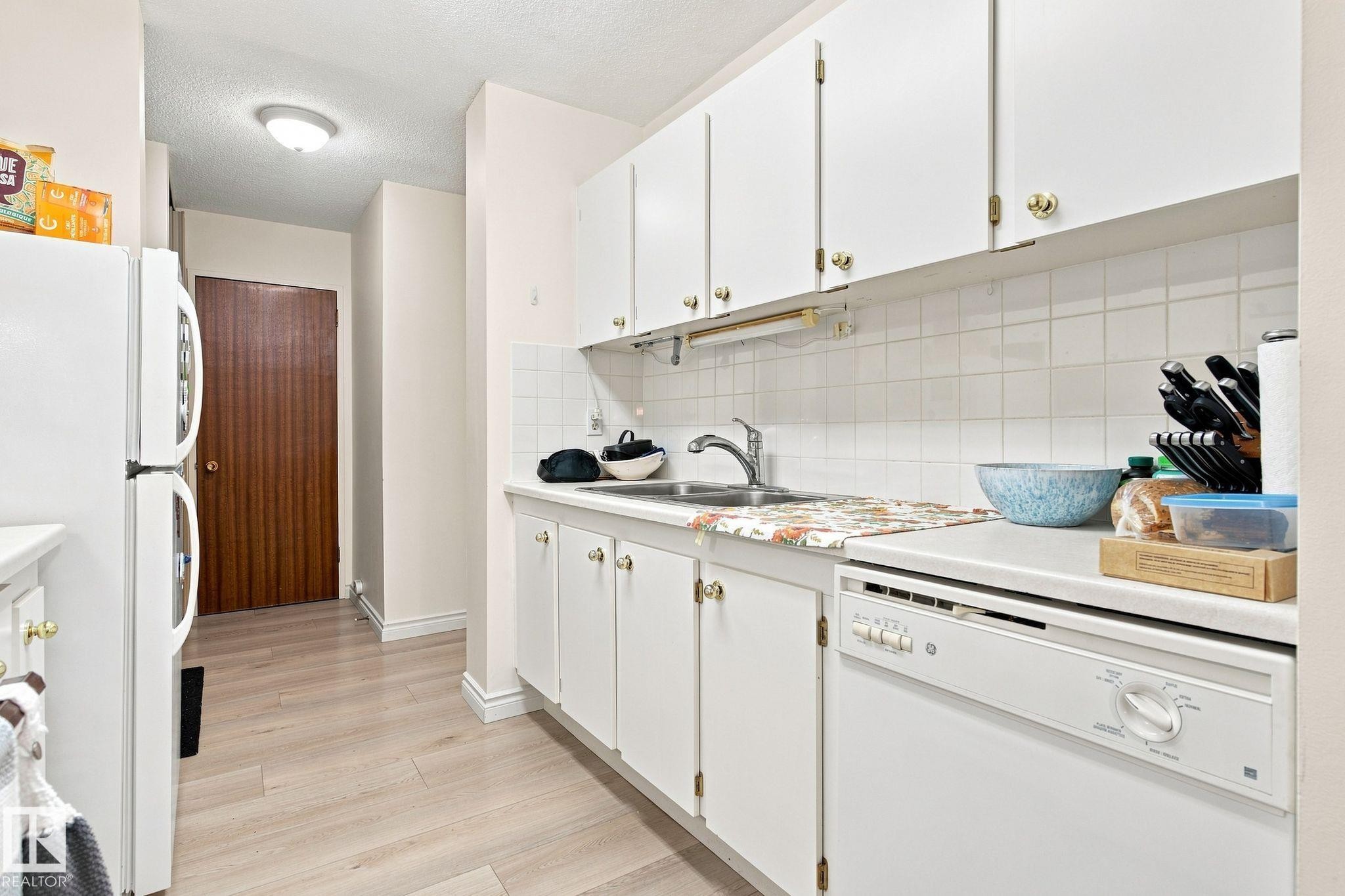 Kitchen featuring white cabinetry with gold-tone hardware, white appliances, light wood-finish flooring, and a tiled backsplash - #1507 9835 113 St Nw, Edmonton, AB
