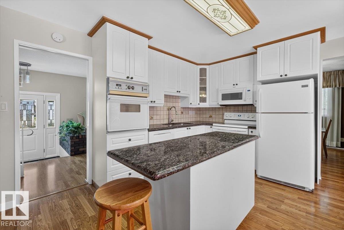 Kitchen featuring white cabinetry with wood trim, built-in appliances, a central island with a granite-finish countertop, and wood-finish flooring - 4027 74 Street Nw, Edmonton, AB - Indoor Photo Showing Kitchen