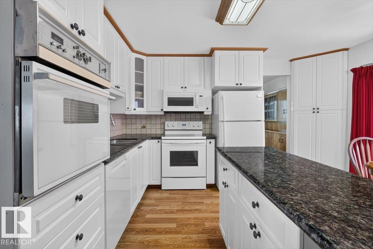 Kitchen featuring white cabinetry with dark hardware, dark granite-look countertops, wood-finish flooring, and white built-in appliances - 4027 74 Street Nw, Edmonton, AB - Indoor Photo Showing Kitchen