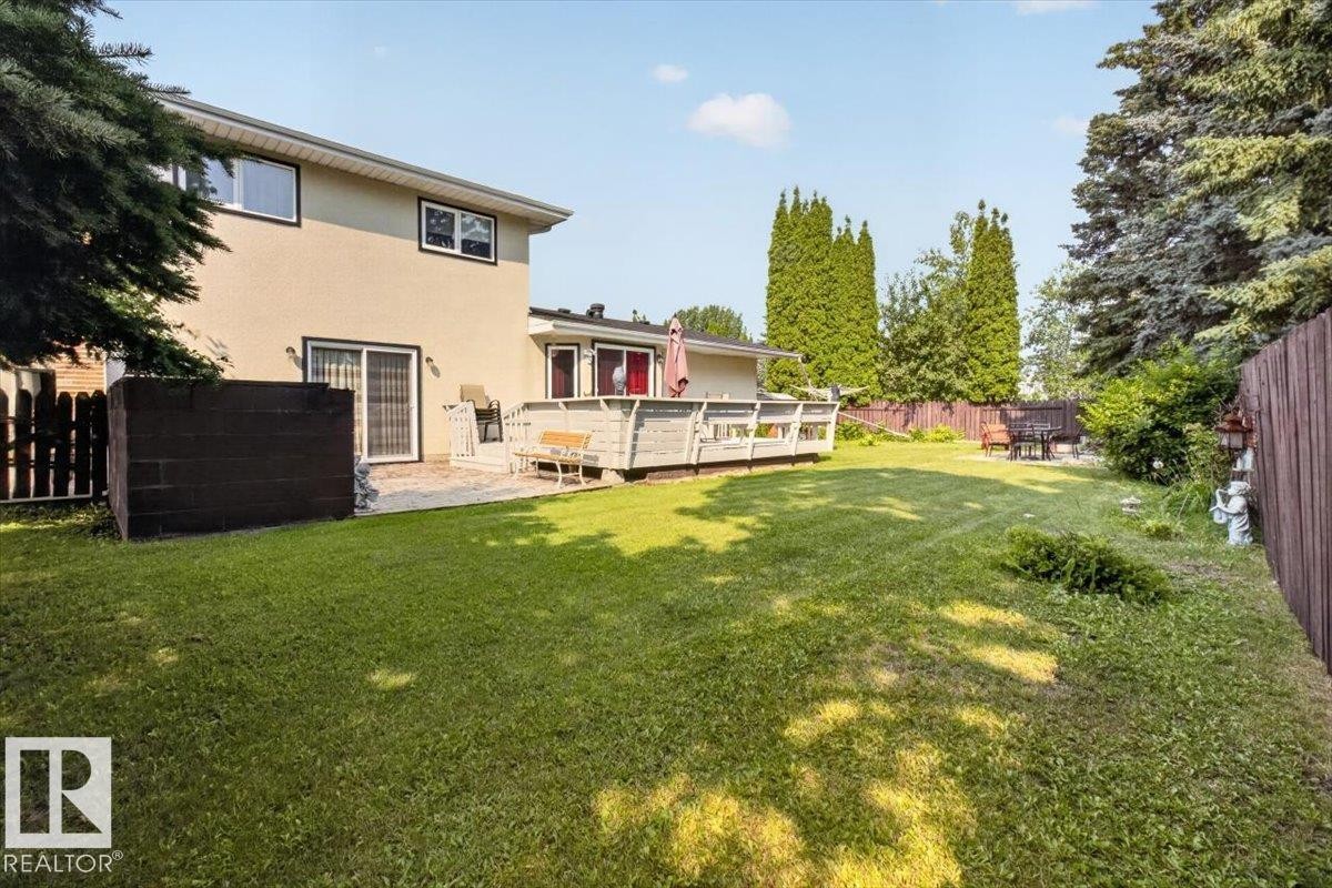 Expansive grassy yard with mature perimeter trees, featuring a raised outdoor deck, a paved patio, and a privacy fence - 4027 74 Street Nw, Edmonton, AB - Outdoor With Deck Patio Veranda
