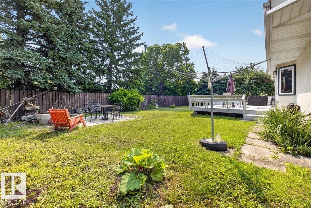 Expansive lawn area featuring a raised wooden deck with built-in bench seating, a separate patio space, a dark wood privacy fence, and mature perimeter trees - 4027 74 Street Nw, Edmonton, AB - Outdoor