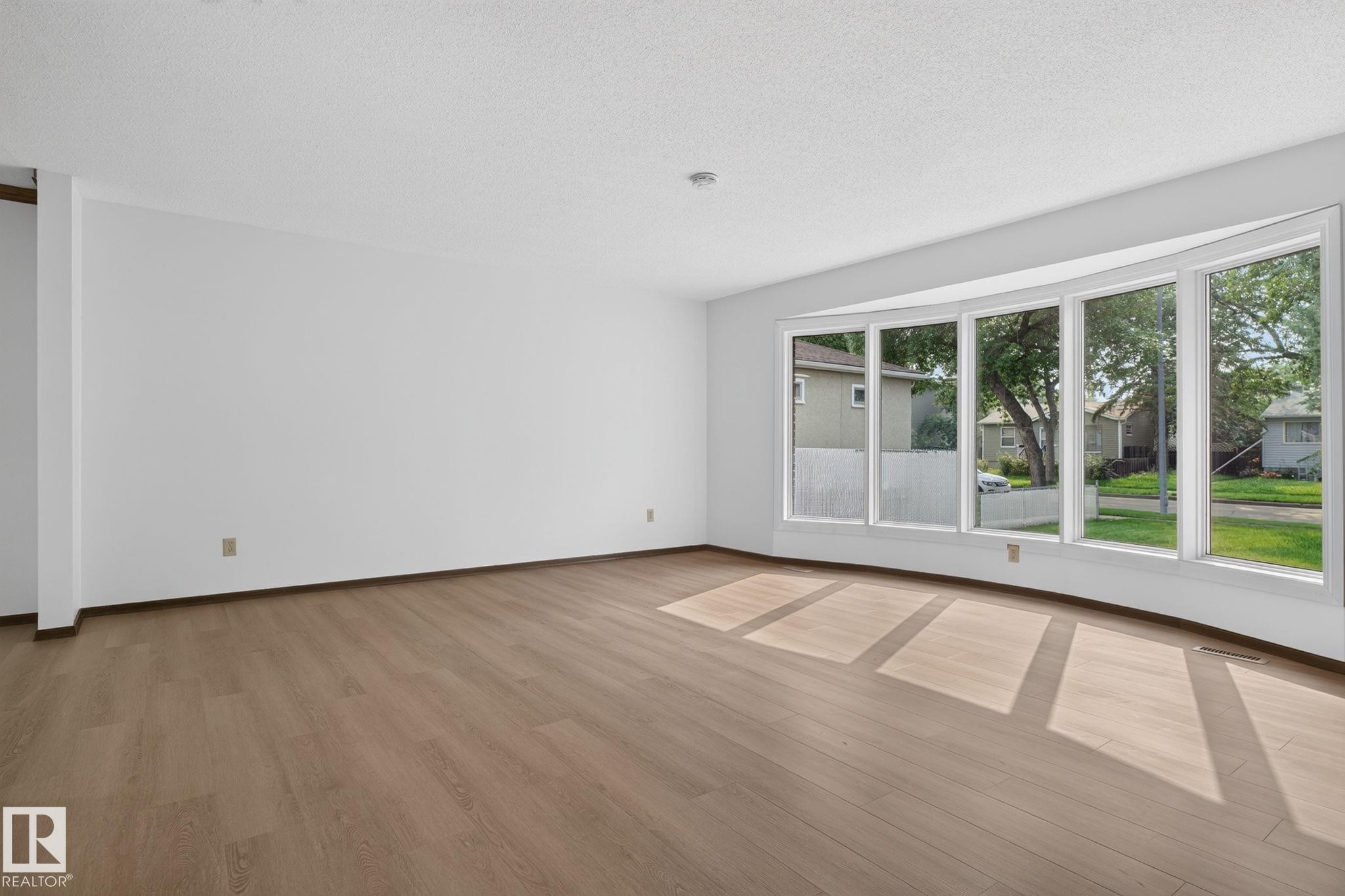 Expansive living space featuring a five-panel bay window, light wood-finish flooring, white walls, and a textured white ceiling - 12121 42 Street Nw, Edmonton, AB - Indoor Photo Showing Other Room