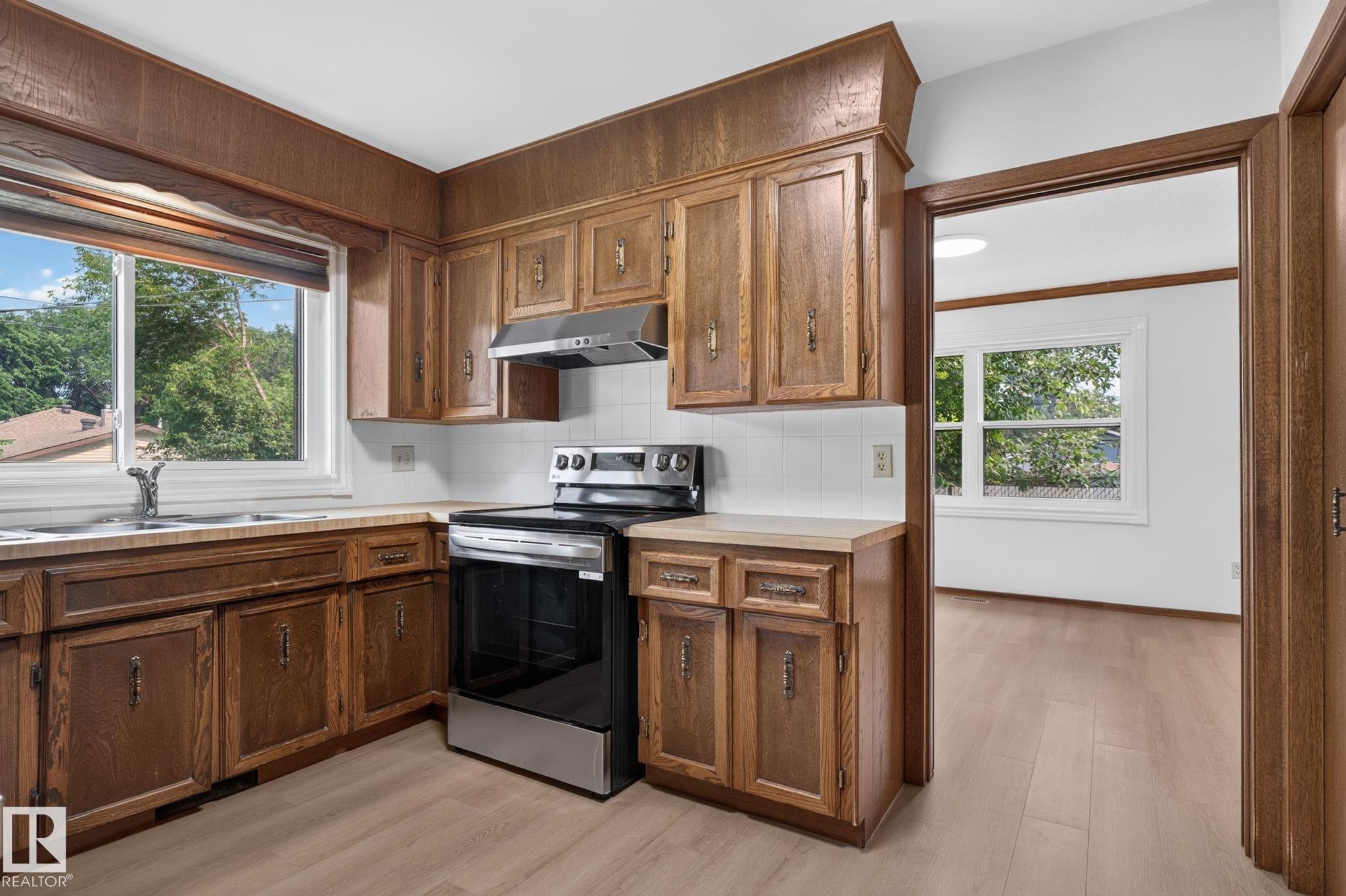 Kitchen featuring wood cabinetry, white tile backsplash, light-toned countertops, and wood-finish flooring - 12121 42 Street Nw, Edmonton, AB - Indoor Photo Showing Kitchen With Double Sink