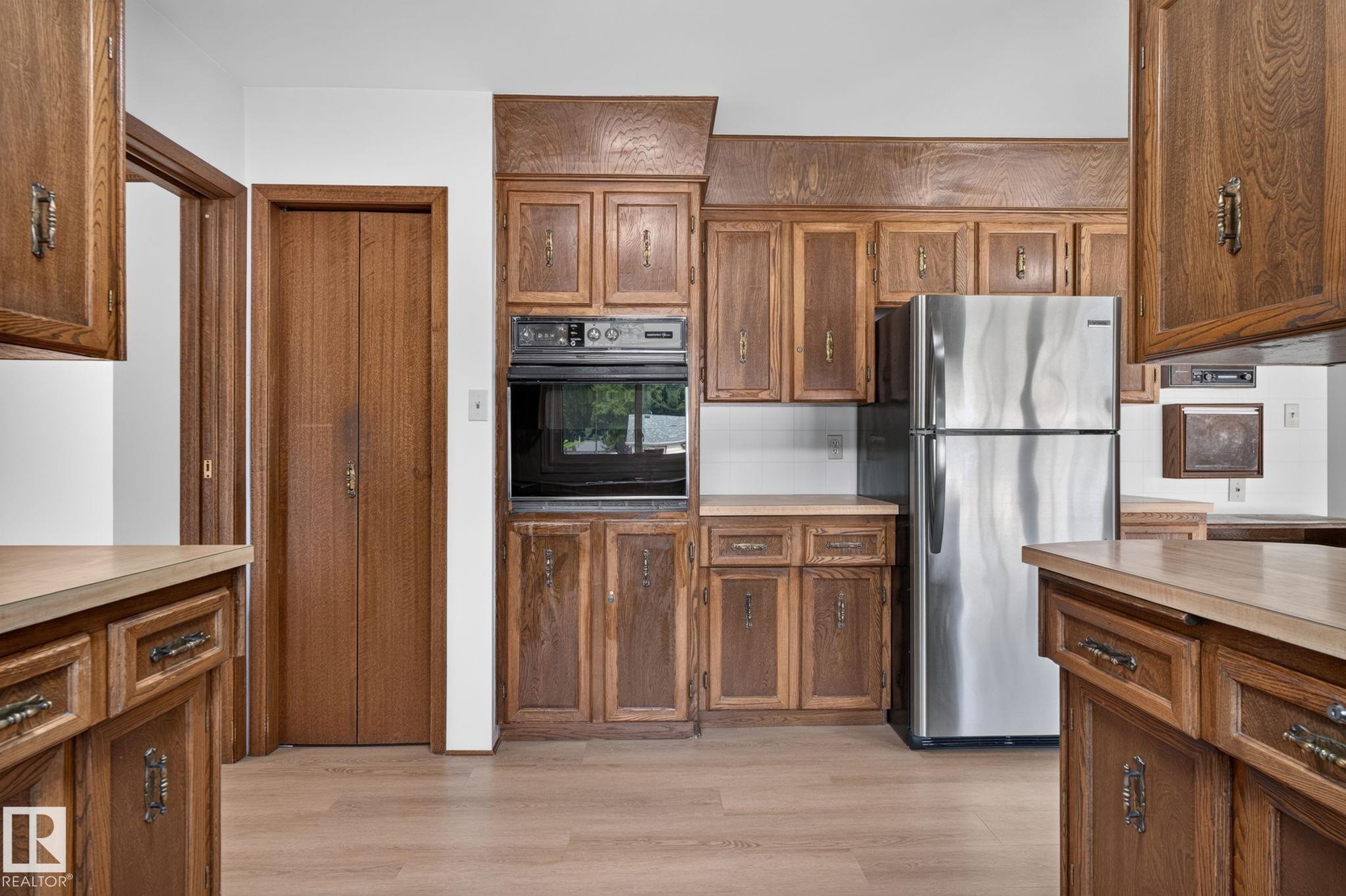 Kitchen featuring wood cabinetry, stainless steel refrigerator, built-in oven, laminate countertops, and wood-finish flooring - 12121 42 Street Nw, Edmonton, AB - Indoor Photo Showing Kitchen