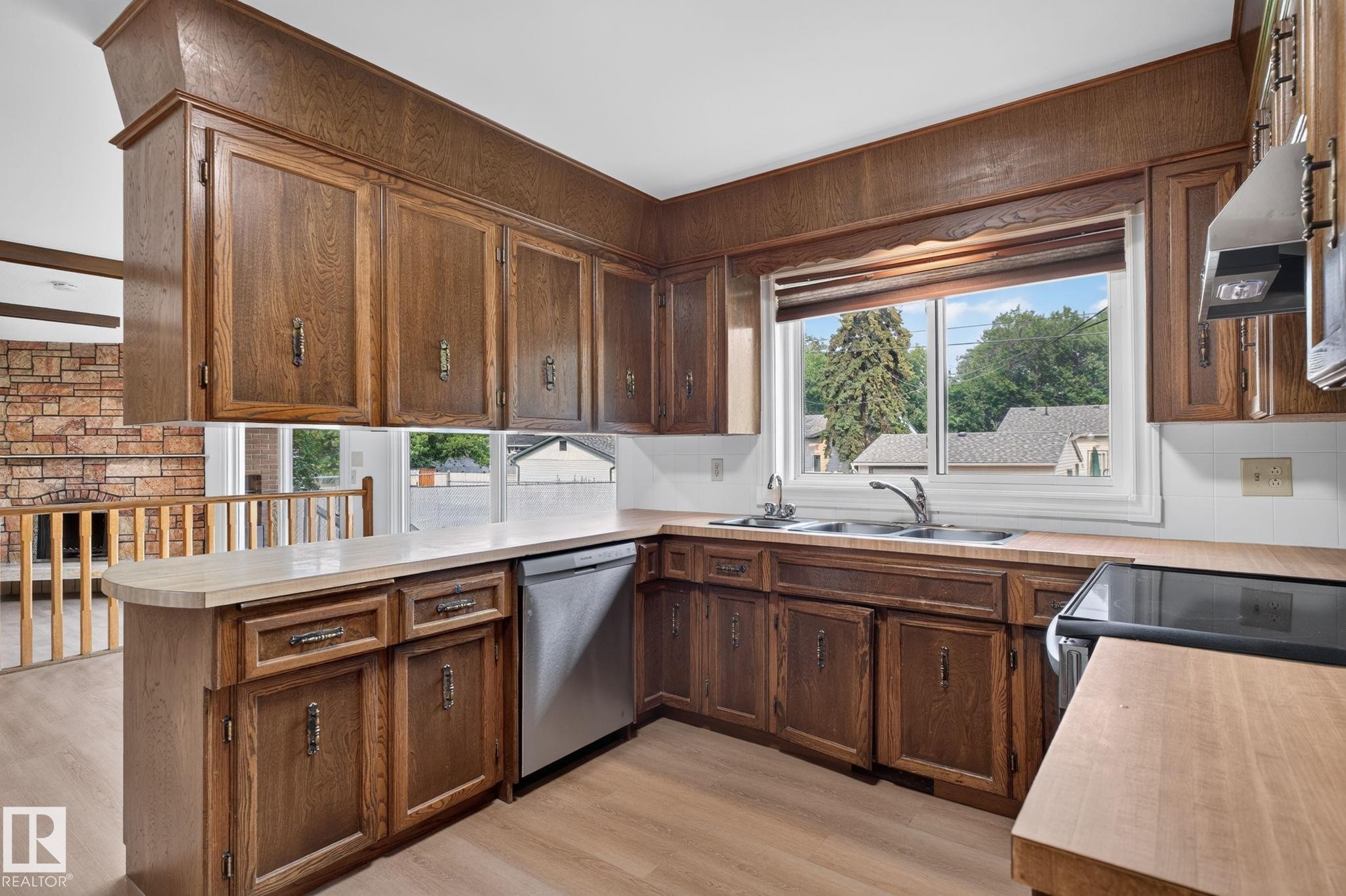 Corner kitchen featuring wood cabinetry, light-toned countertops, a double basin sink with a window, and a stainless steel dishwasher - 12121 42 Street Nw, Edmonton, AB - Indoor Photo Showing Kitchen With Double Sink