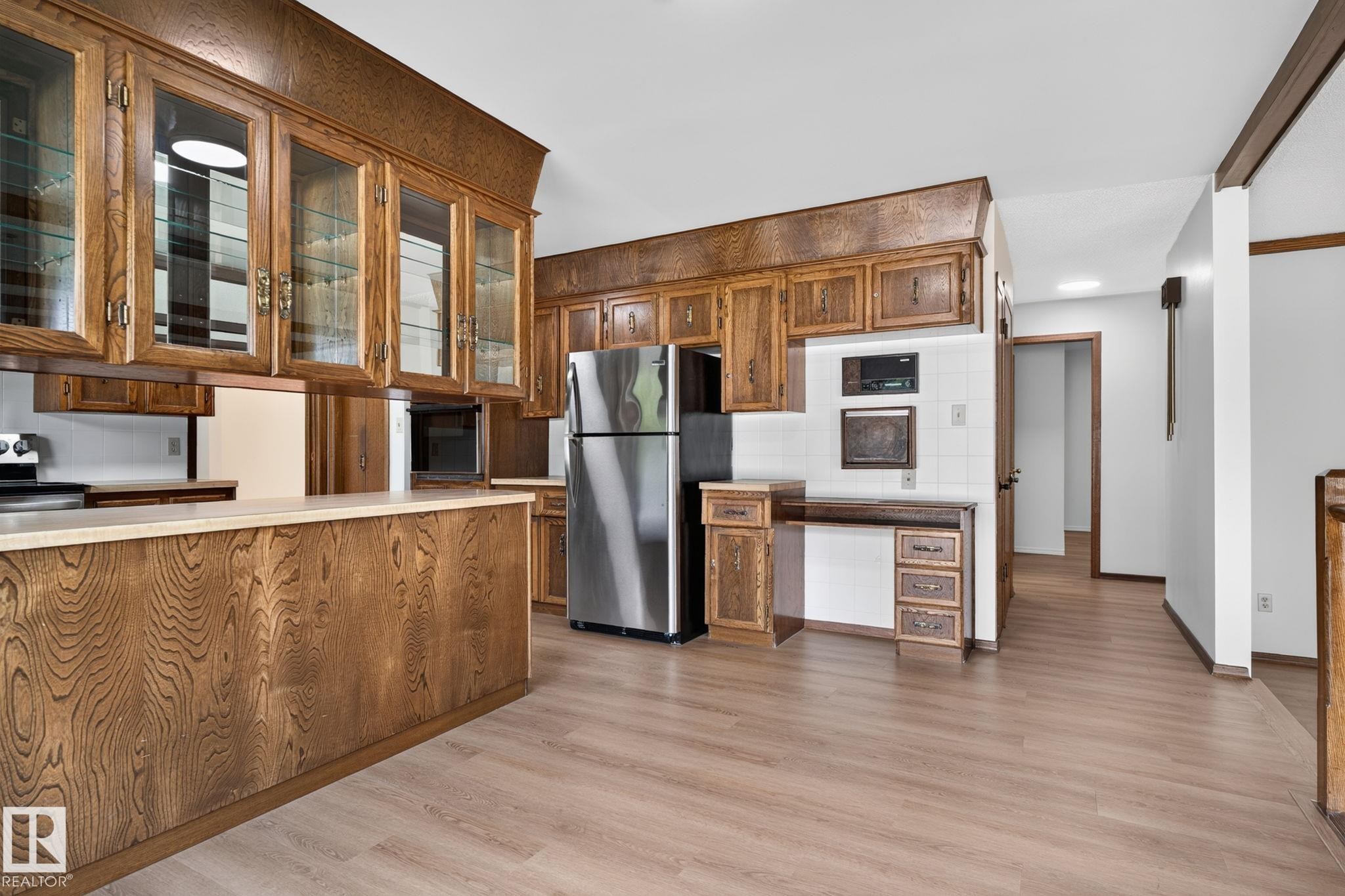 Kitchen featuring wood-finish cabinetry, integrated glass-front display cabinets, light-toned countertops, tile backsplash, and wood-finish flooring - 12121 42 Street Nw, Edmonton, AB - Indoor Photo Showing Kitchen With Stainless Steel Kitchen