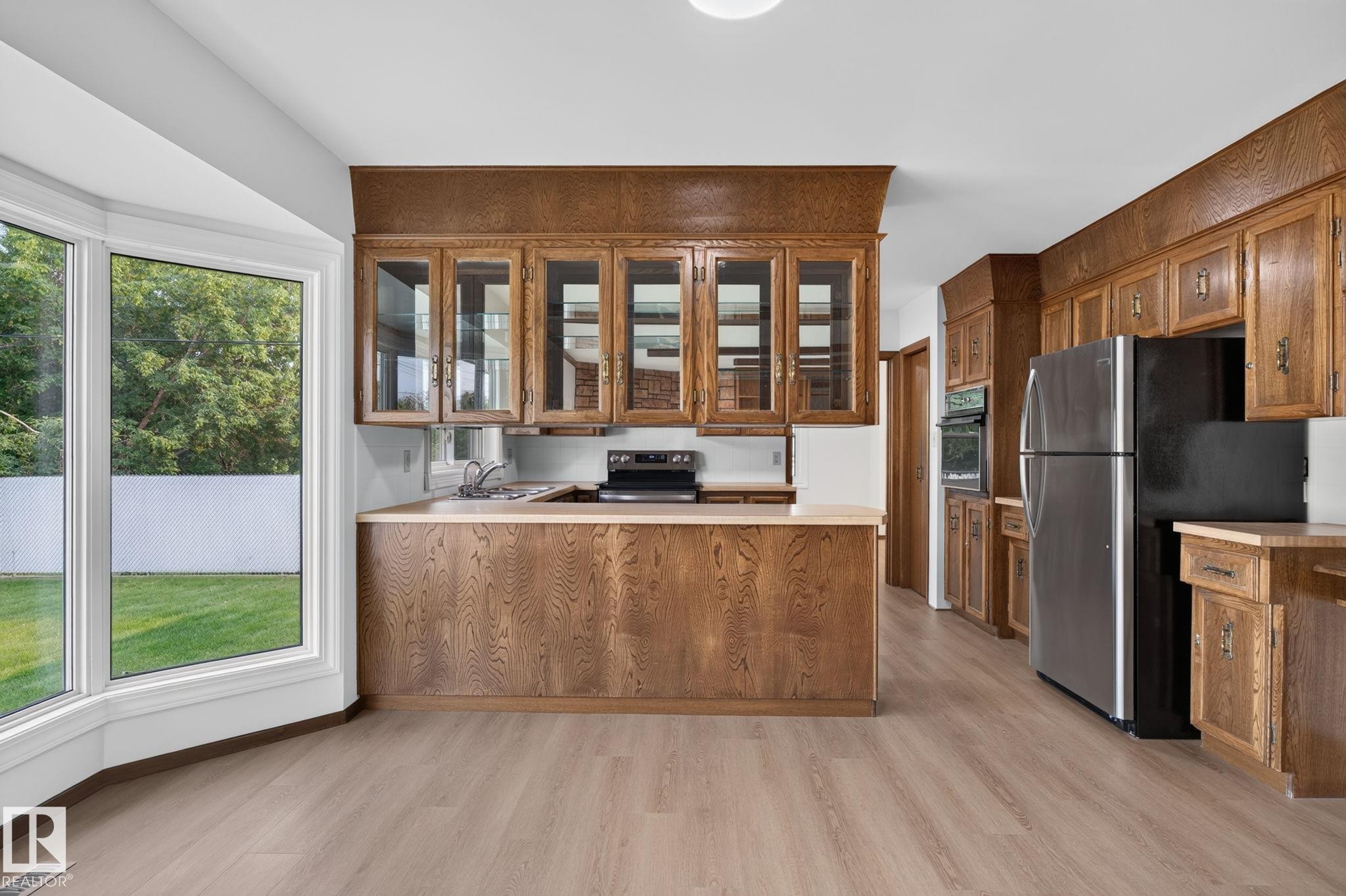 Kitchen featuring extensive wood cabinetry, a bay window overlooking a green space, stainless steel refrigerator, built-in wall oven, and light wood-finish flooring - 12121 42 Street Nw, Edmonton, AB - Indoor Photo Showing Kitchen