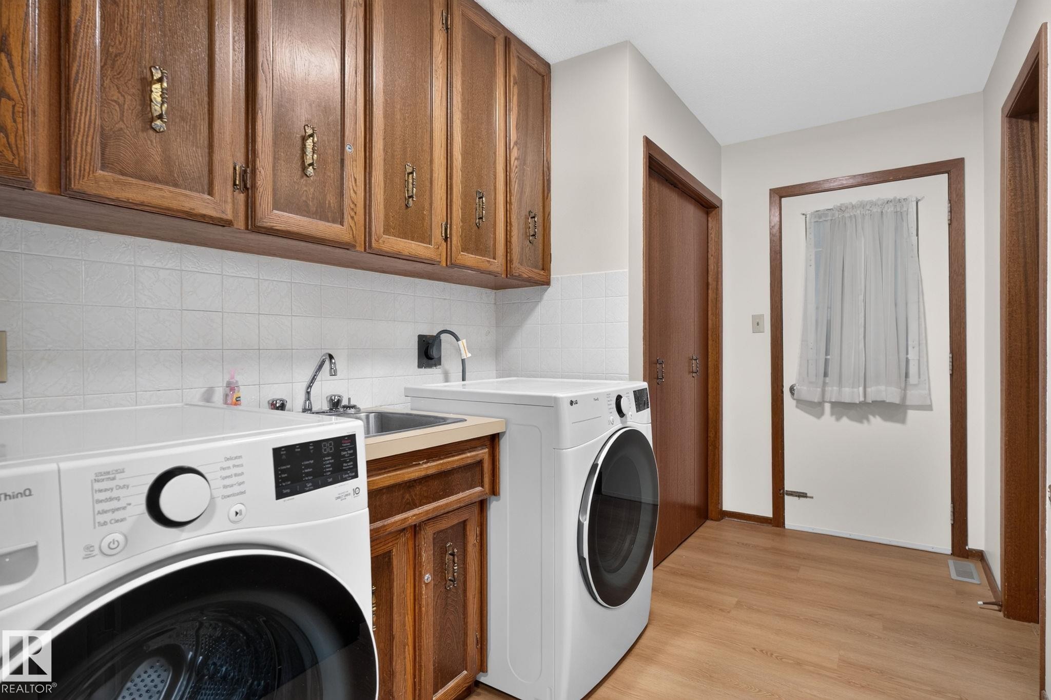 Dedicated laundry room featuring wood-finish flooring, wood cabinetry with decorative hardware, a utility sink, and white tile backsplash - 12121 42 Street Nw, Edmonton, AB - Indoor Photo Showing Laundry Room