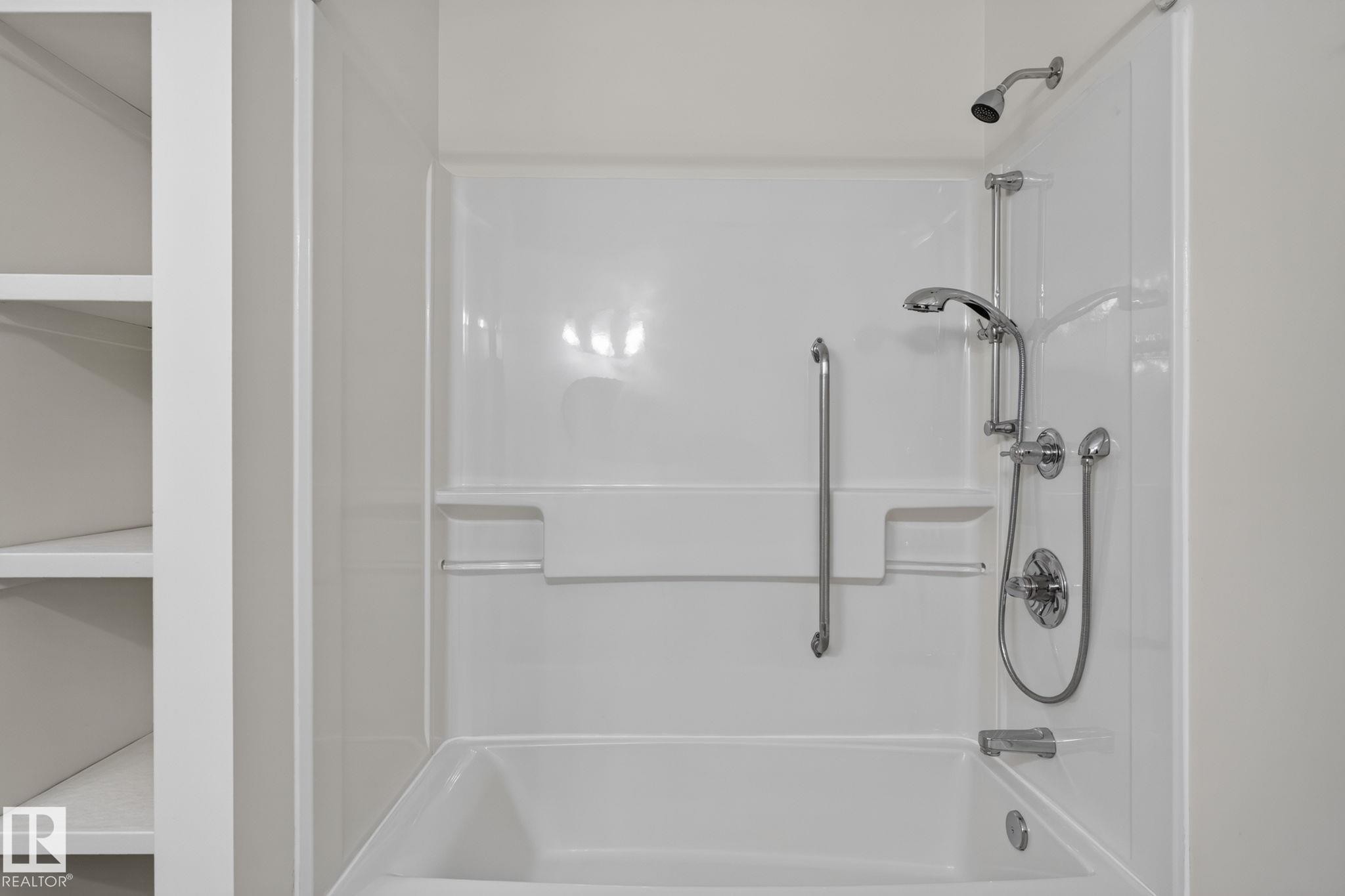 Bathtub and shower combination featuring a multi-function showerhead with a handheld spray, integrated shelving, and a grab bar - 12121 42 Street Nw, Edmonton, AB - Indoor Photo Showing Bathroom