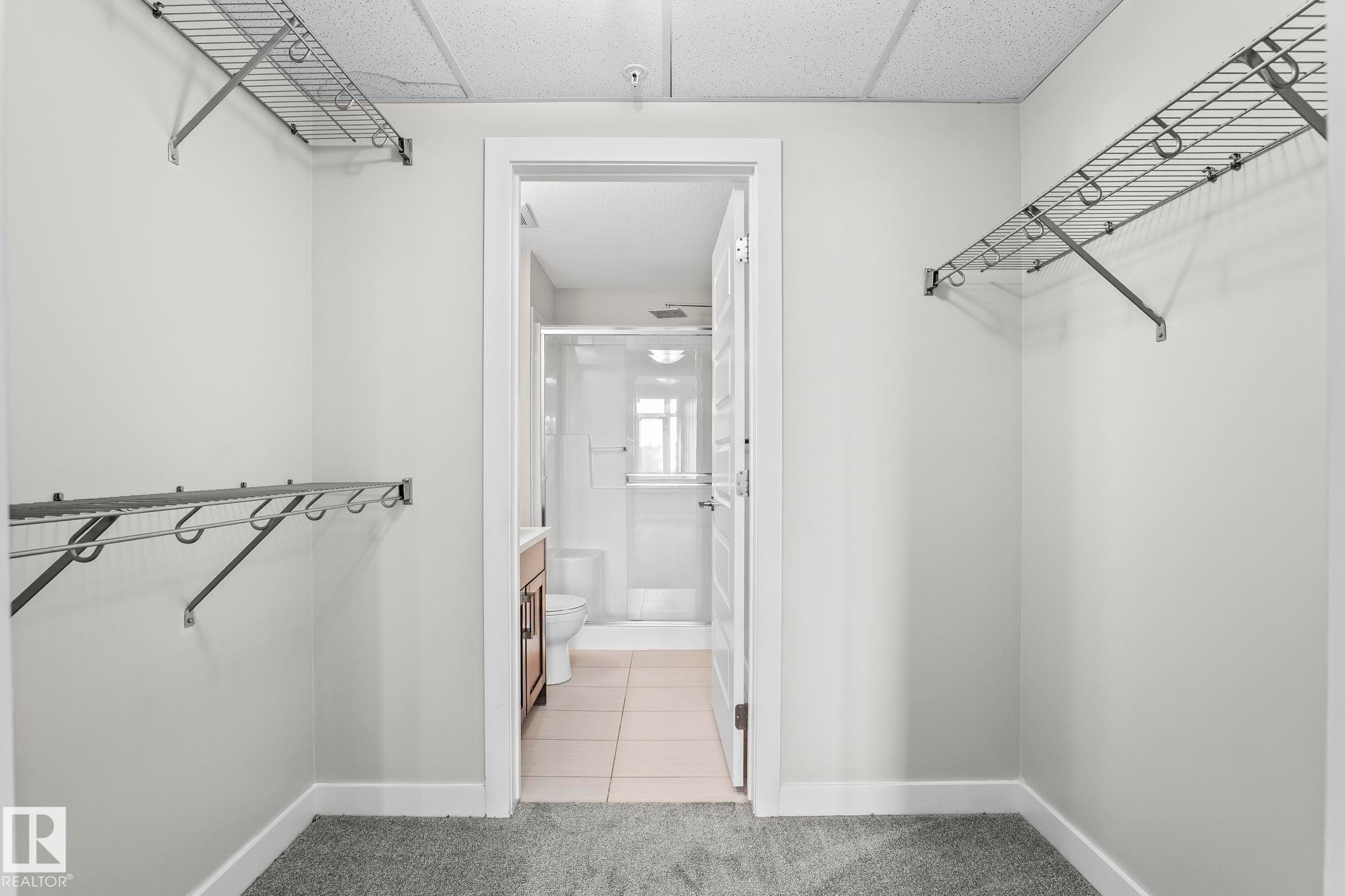 Walk-in closet with carpet flooring, wire shelving, and direct access to a bathroom featuring a glass-enclosed shower, a vanity with wood-finish cabinetry, and tile flooring - #324 5151 Windermere Bv Sw, Edmonton, AB - Indoor With Storage
