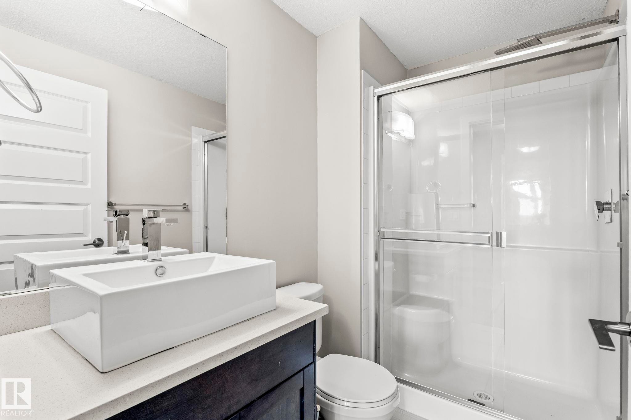 Modern bathroom featuring a rectangular vessel sink with a chrome faucet, light-toned countertop, dark wood-finish vanity, and a glass-enclosed shower with a sliding door - #324 5151 Windermere Bv Sw, Edmonton, AB - Indoor Photo Showing Bathroom