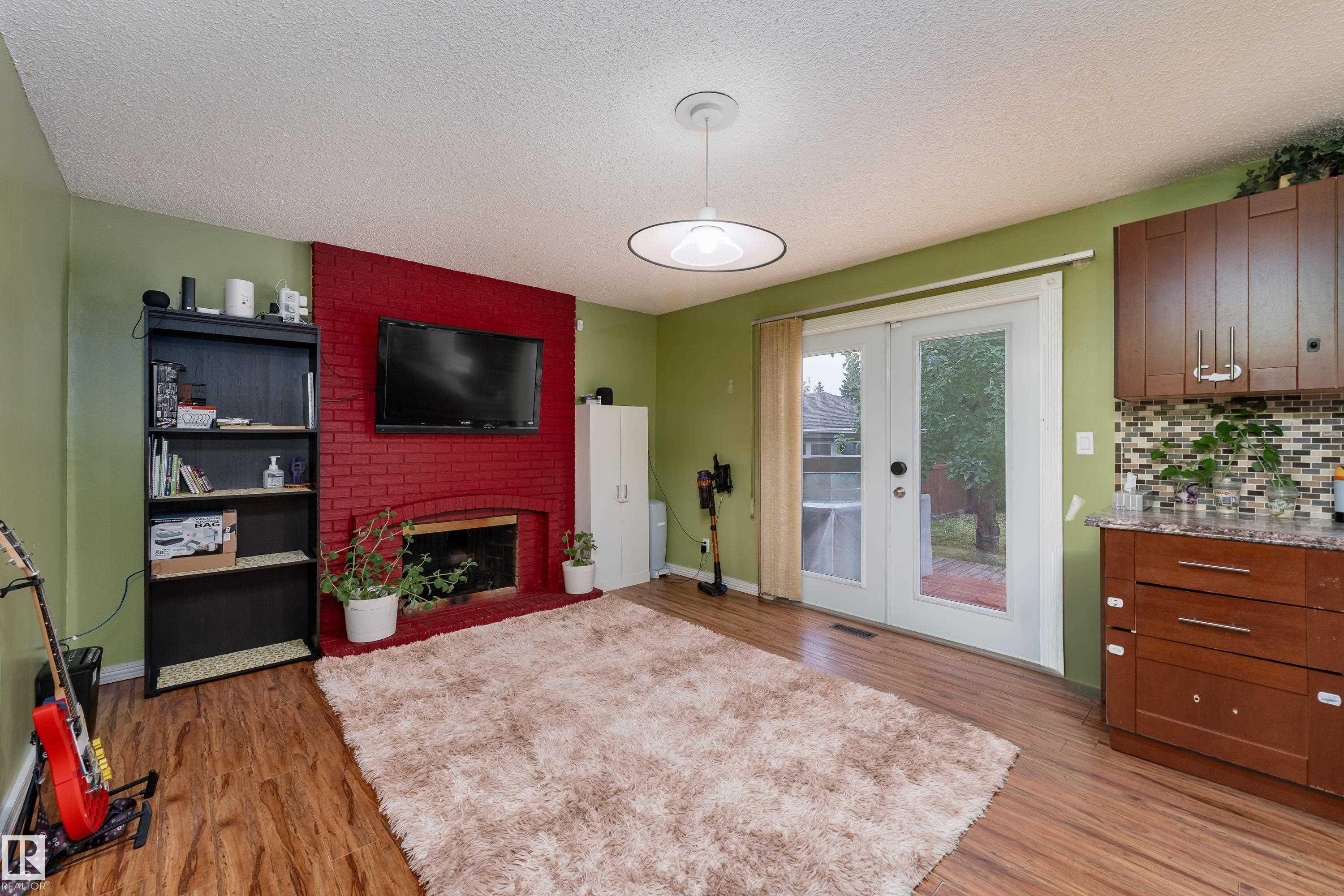 Wood-finish flooring throughout, red brick fireplace, dual glass-paneled patio doors, built-in brown cabinetry with a patterned backsplash, and a contemporary ceiling fixture - 17928 93 Ave Nw, Edmonton, AB