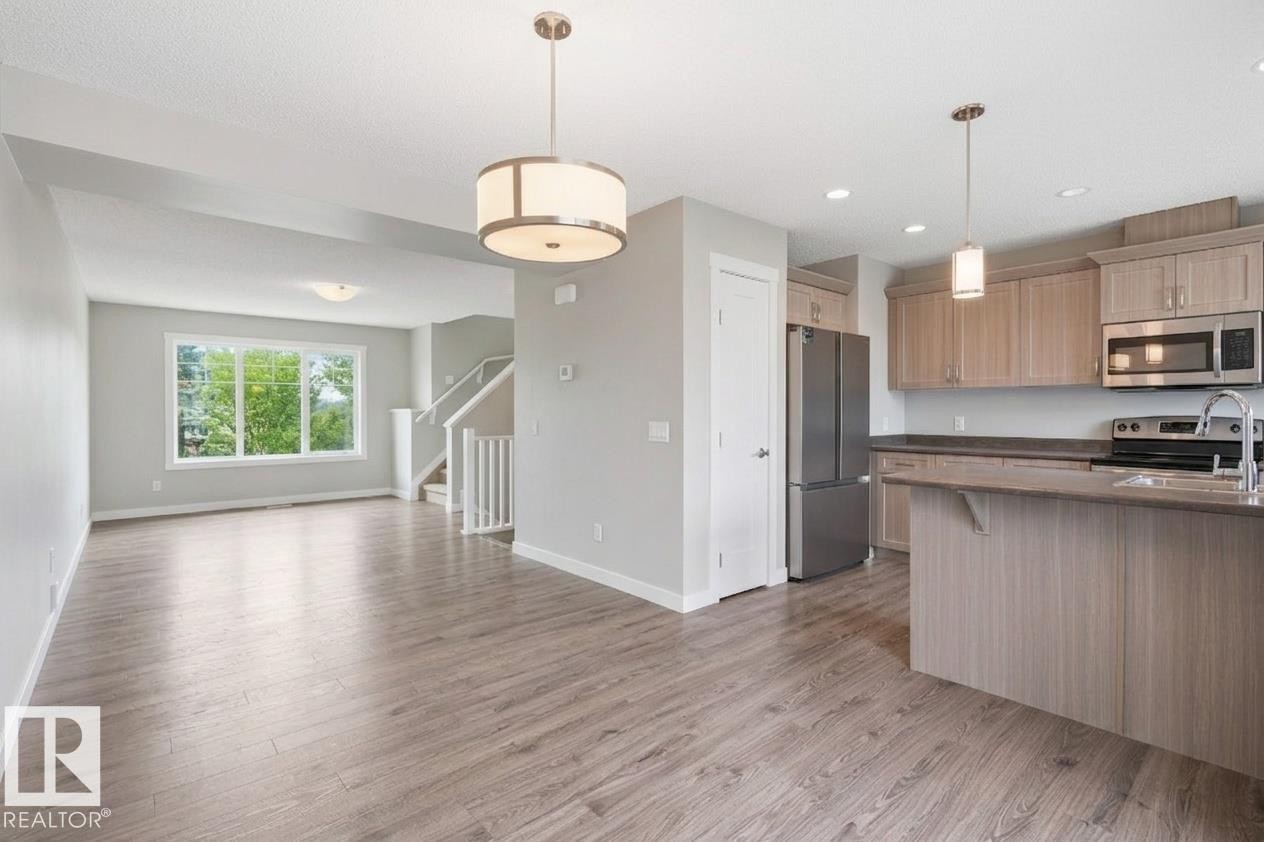 Open-concept living space with wood-finish flooring, a contemporary kitchen, stainless steel appliances, and a central island with a sink - #12 1391 Starling Dr Nw, Edmonton, AB